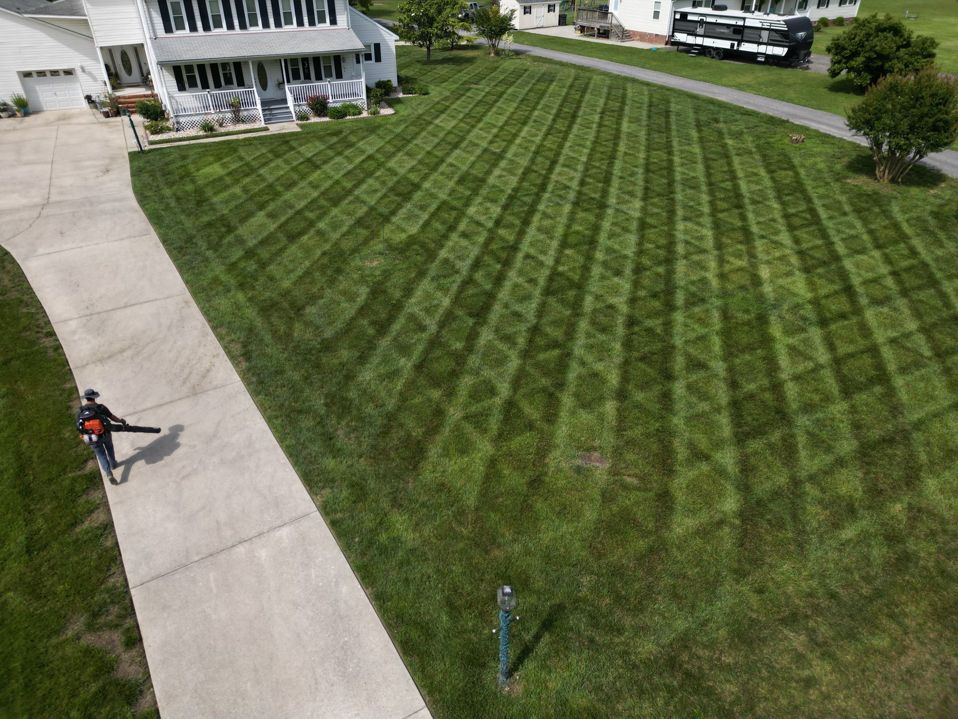 Person using leaf blower on a lawn with a diamond-patterned mowing design next to a sidewalk and house.