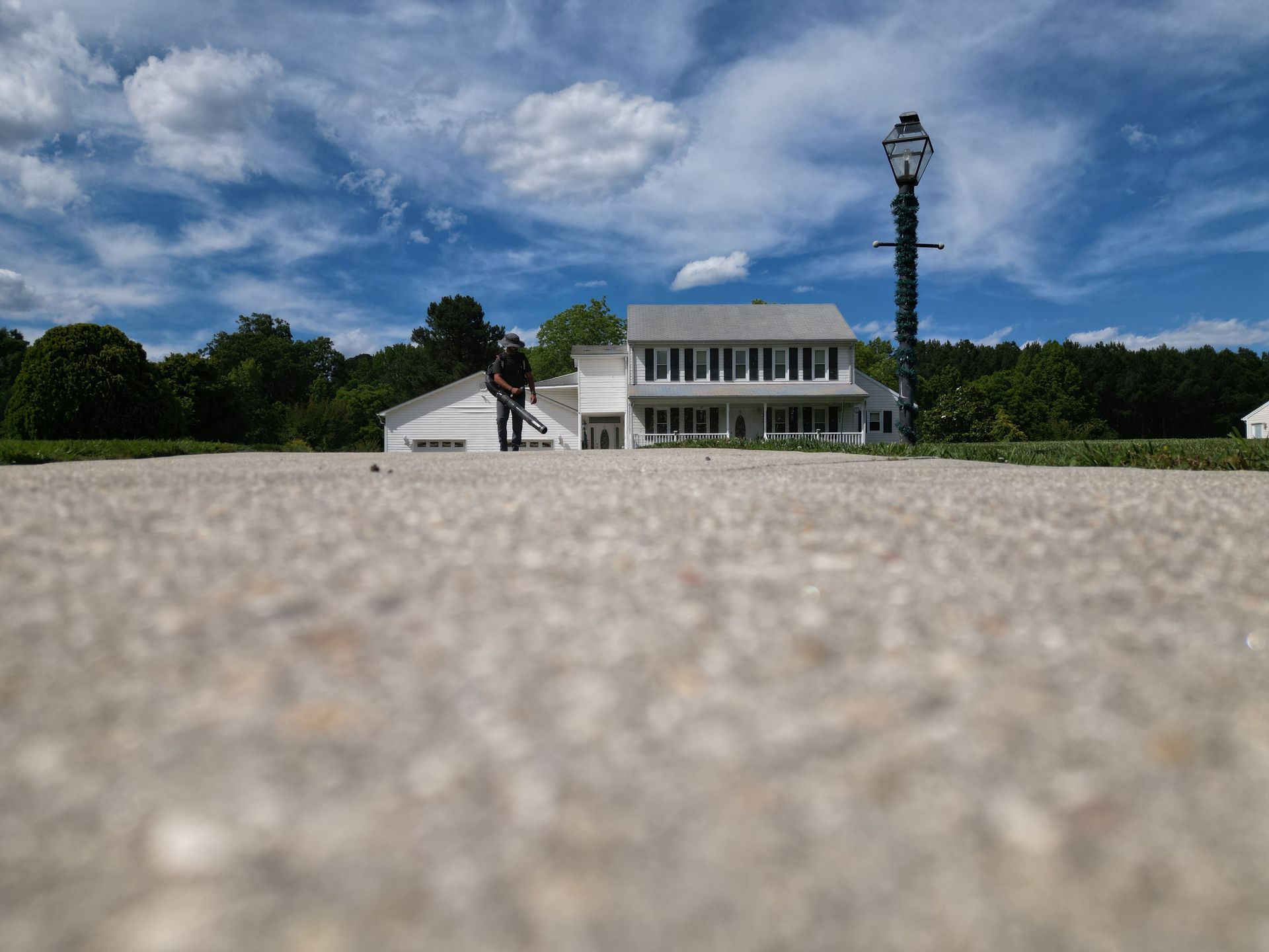 Person mowing grass in front of a two-story house with a cloudy blue sky in the background.