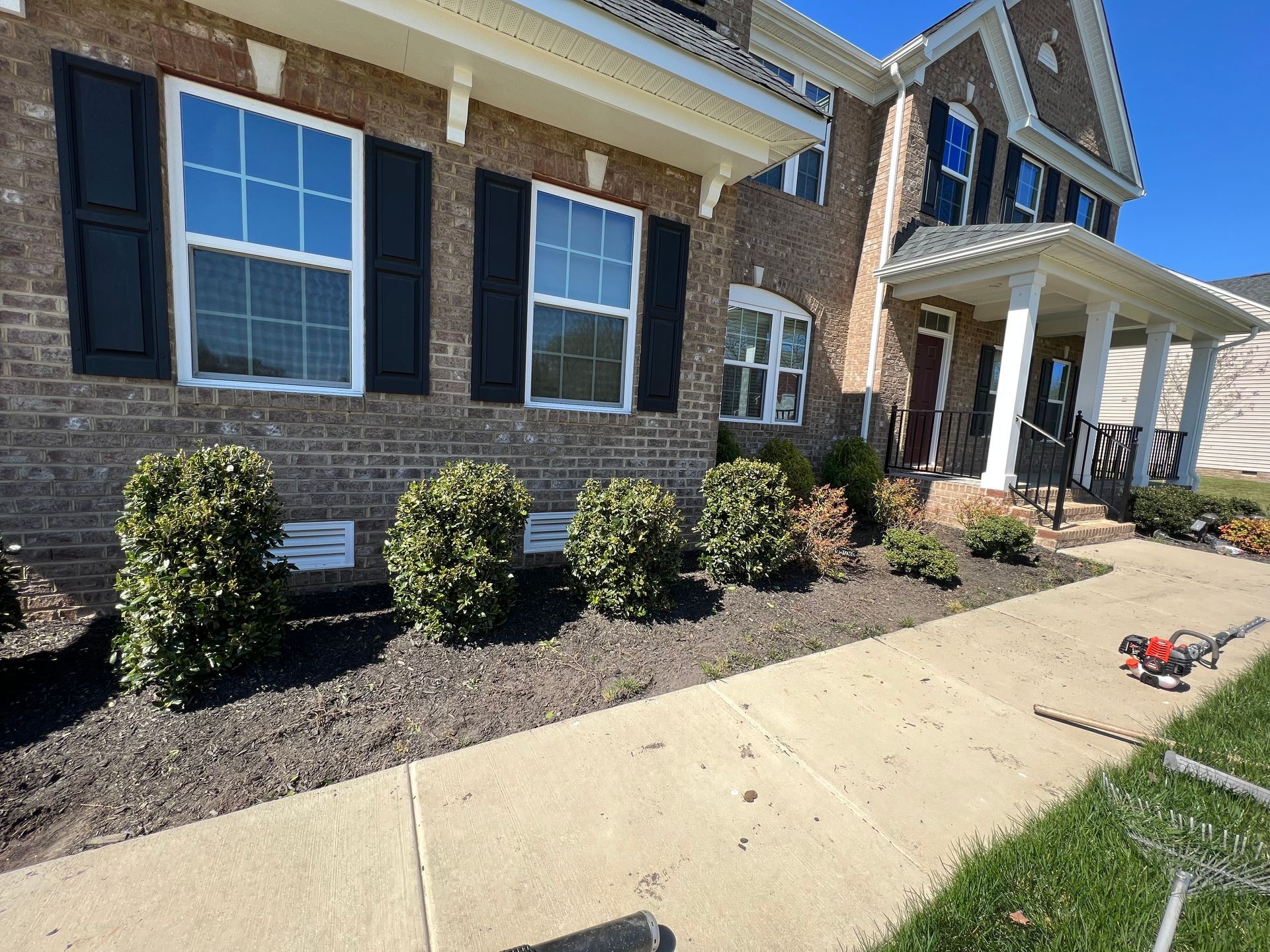 Brick house with black shutters, white trim, and trimmed bushes along a sidewalk.