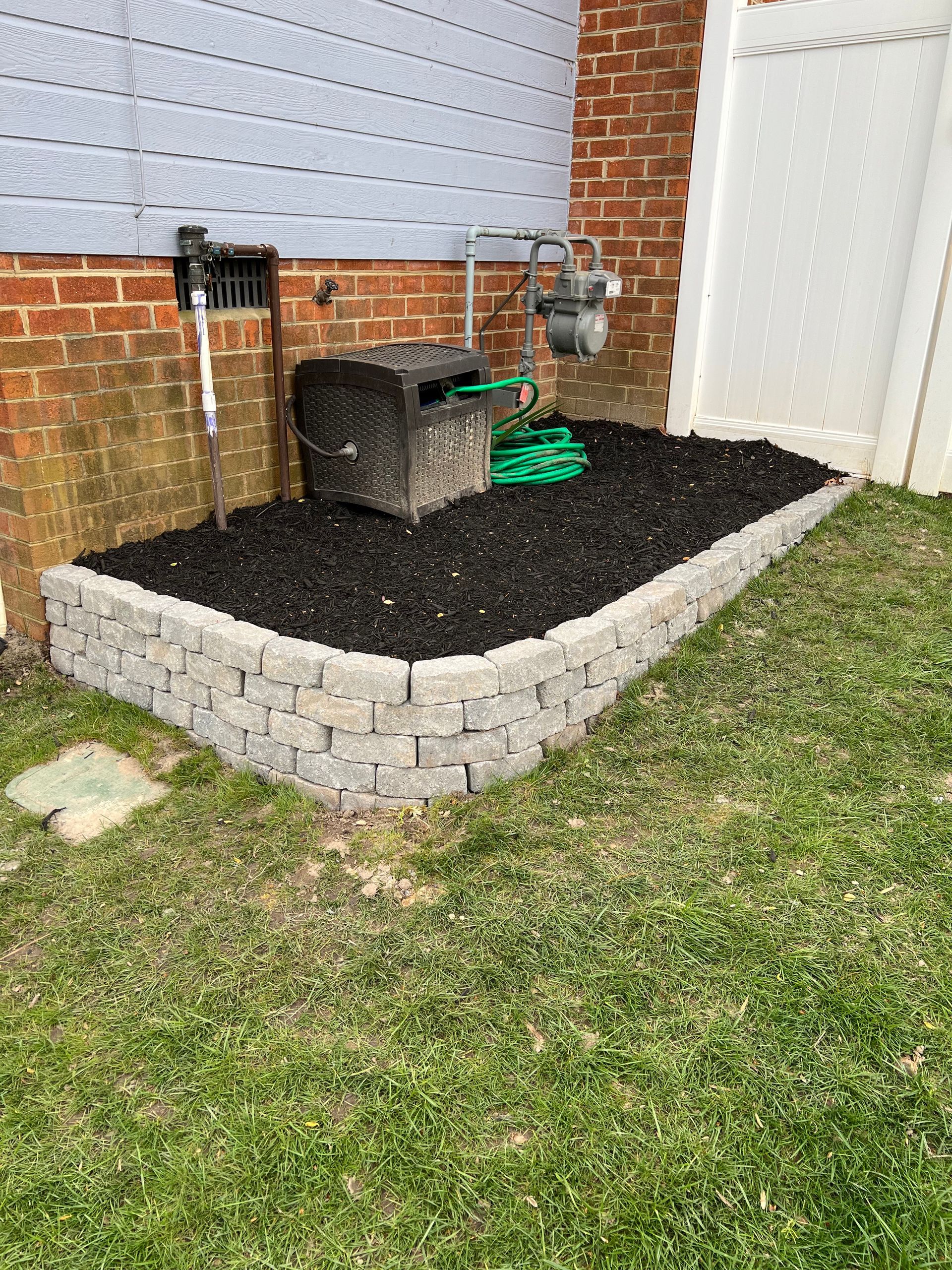 Stone retaining wall with dark mulch bed, beside brick wall, gas meter, and white fence.