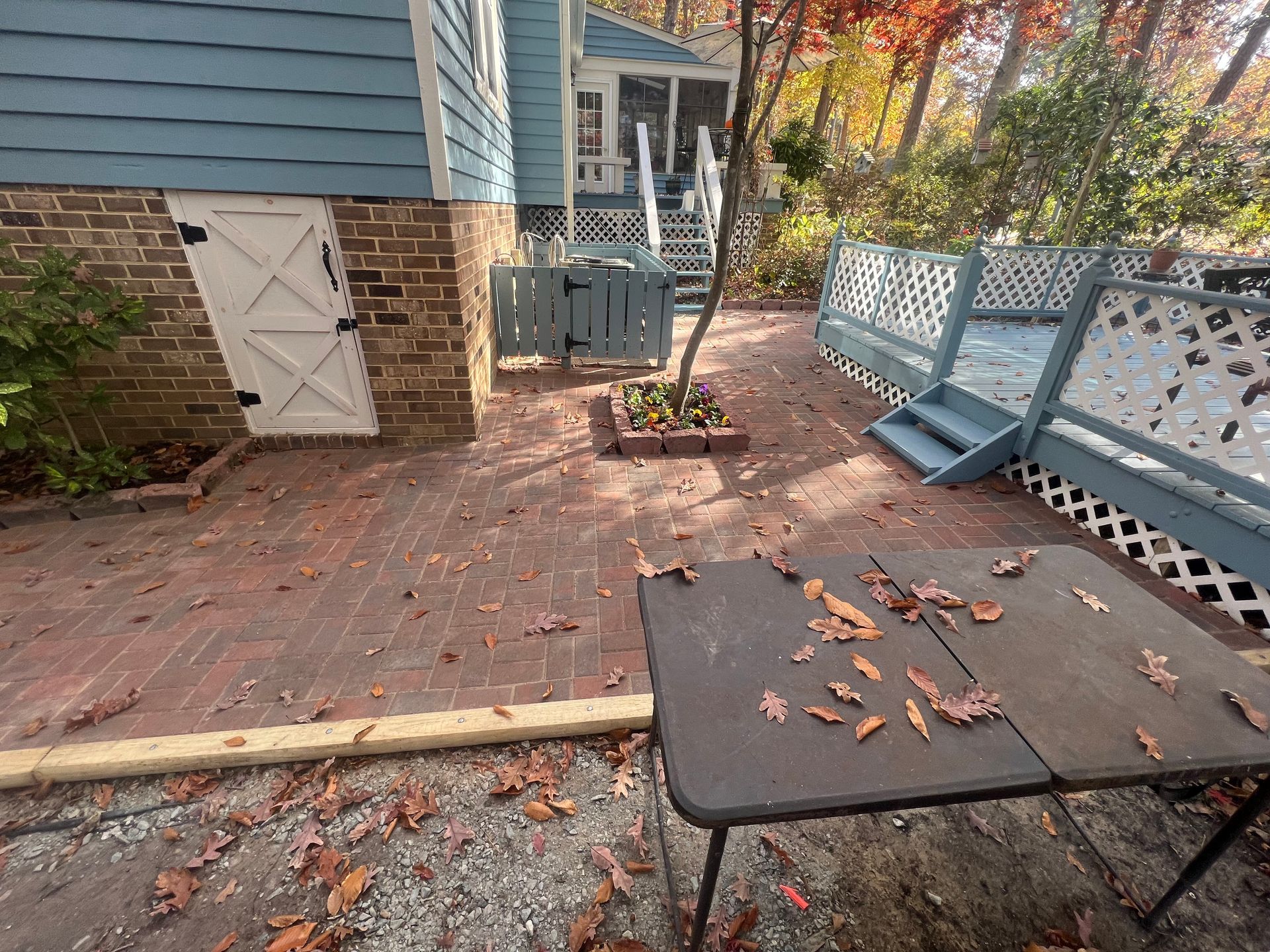 Brick patio with blue house and deck, brown table with leaves, autumn trees in the background.