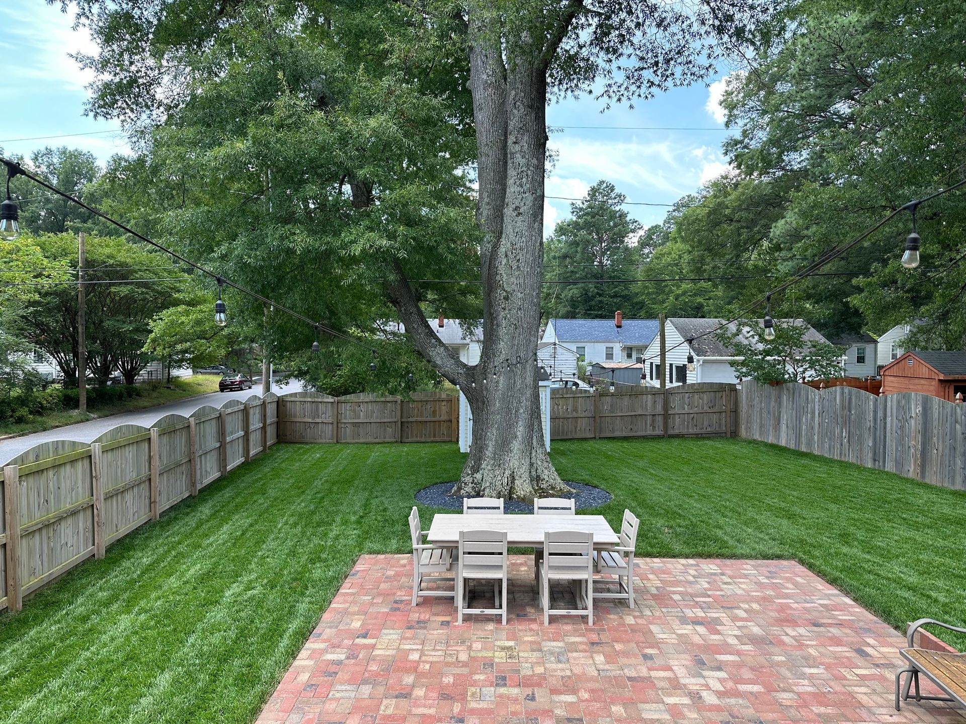 A backyard with a brick patio, table and chairs, and a large tree surrounded by green grass and a wooden fence.