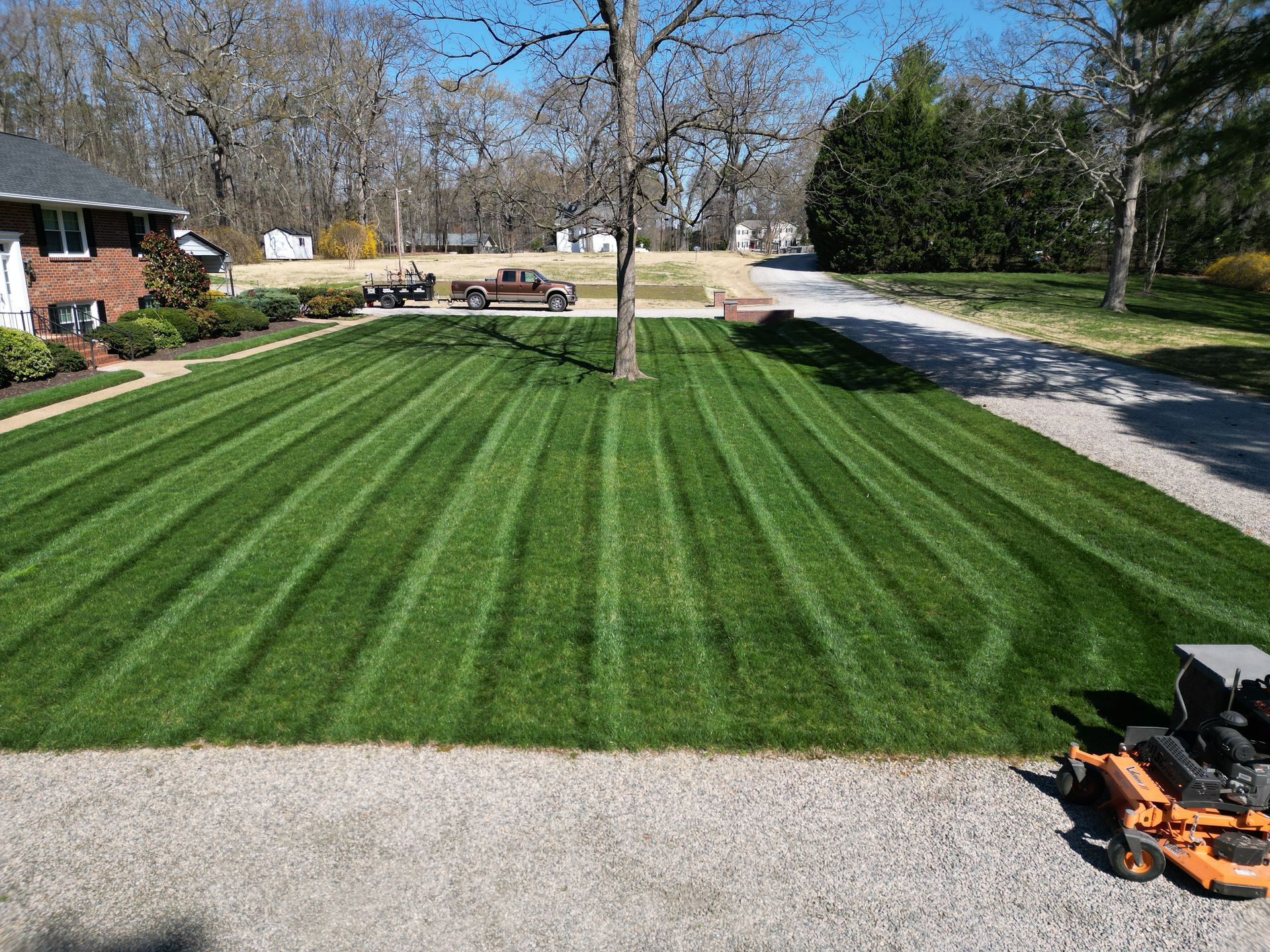 Lush green lawn with distinct mowing stripes surrounding a tree. Brownstone house and gravel driveway. Bright sunny day.