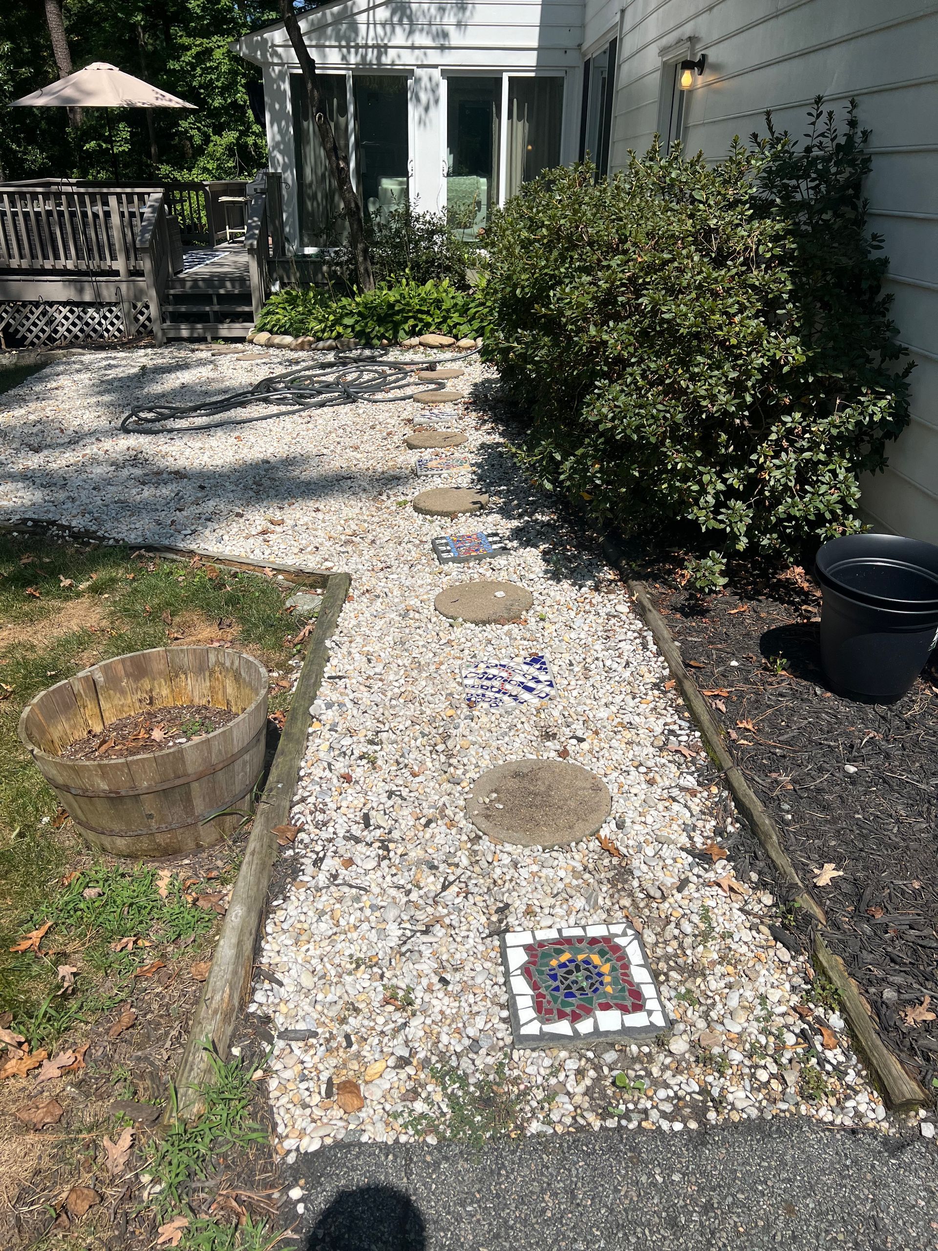 Gravel pathway with stepping stones and mosaic tiles leading to a house entrance. Wooden edges and a bush are nearby.