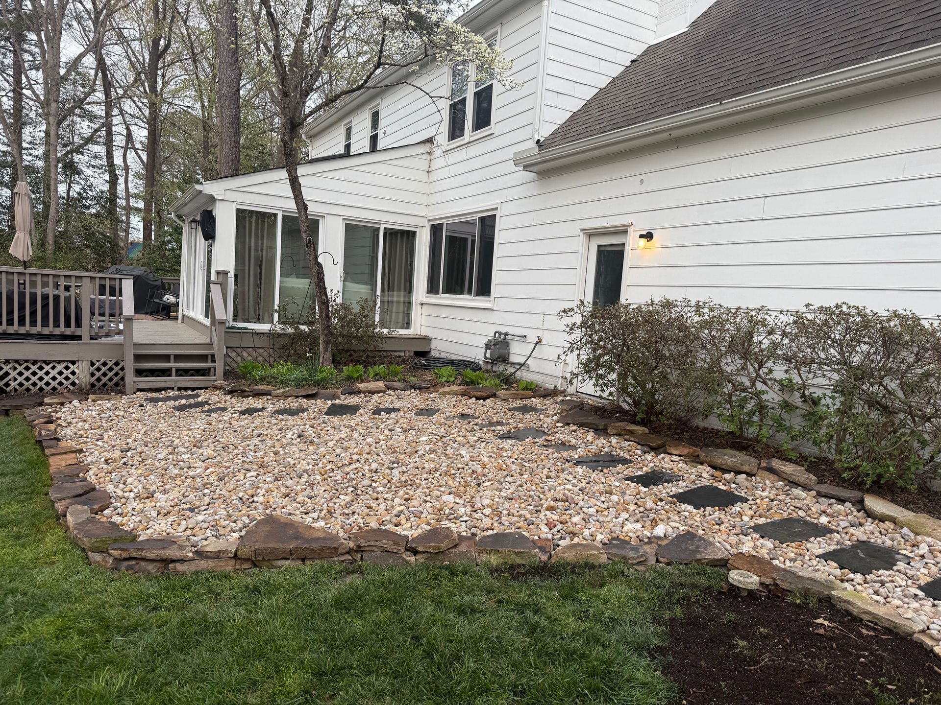 A backyard with a gravel patio, stone pathway, and deck next to a two-story white house.