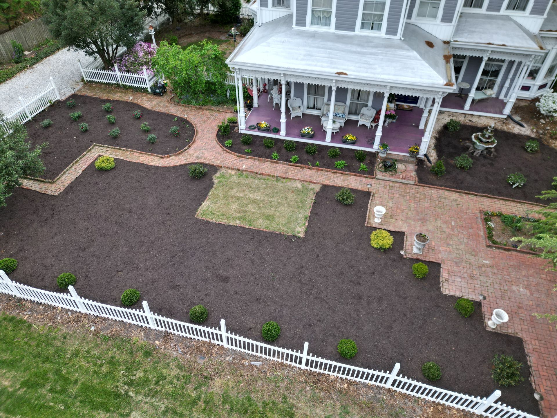 Aerial view of a Victorian house with a landscaped yard. Dark mulch, brick walkways, and a white picket fence are visible.