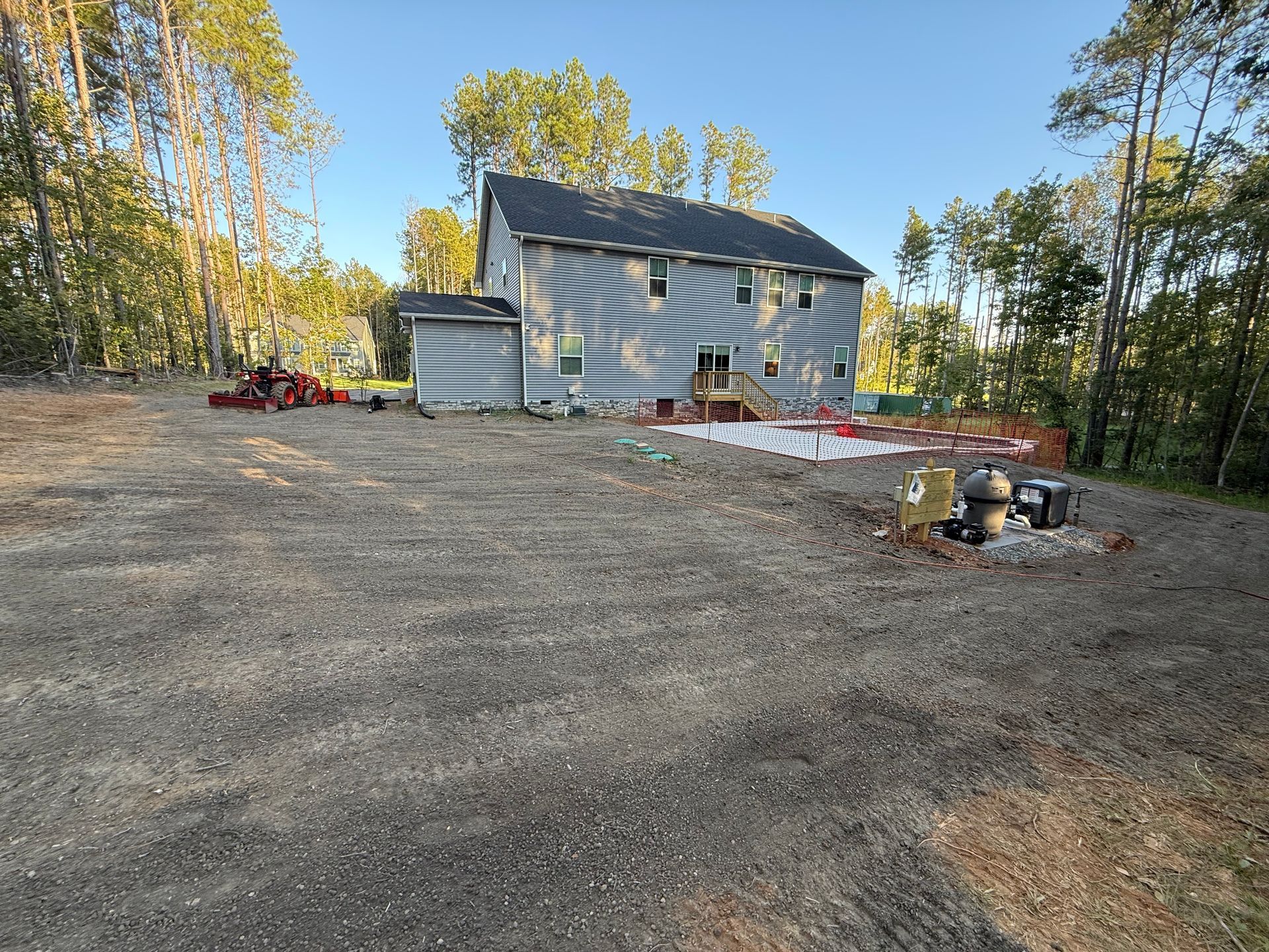 Gravel lot with a two-story house in the background. Construction materials and safety fencing visible. Trees in the distance.