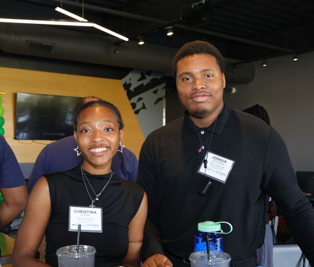 Two young Black adults smile at the camera, standing behind a counter with drinks. They are indoors with a modern design.