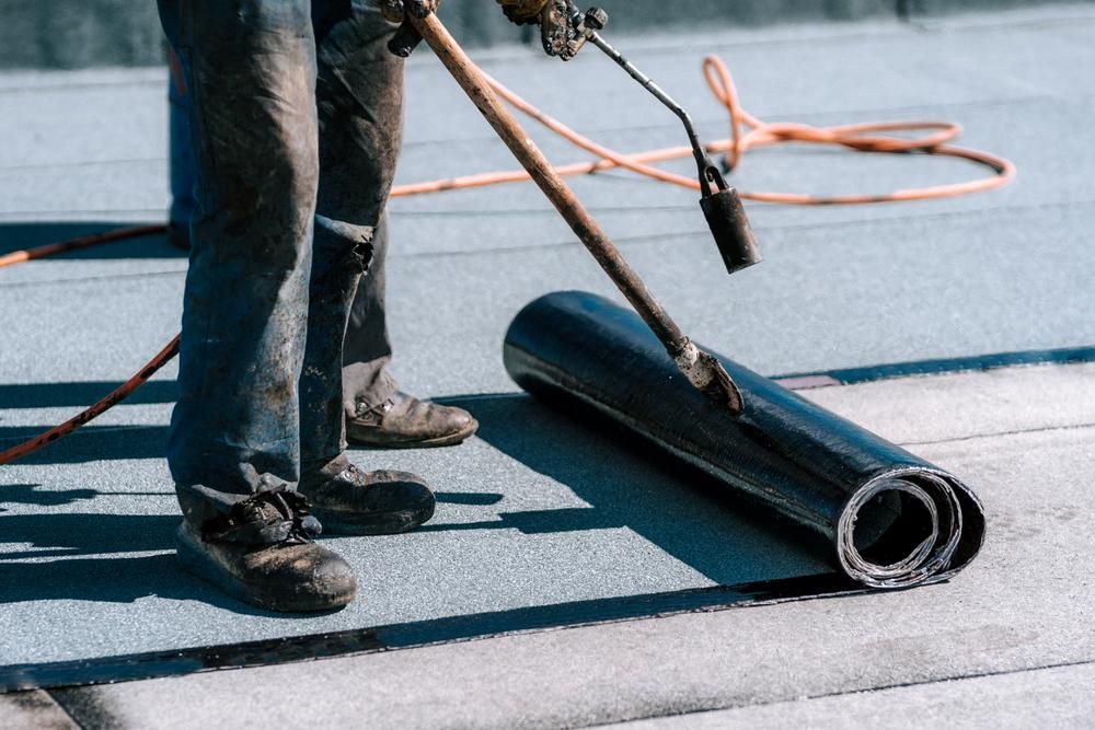 A Man Is Rolling a Roll of Roofing Material on A Roof — John Chisholm Flooring in Mareeba, QLD