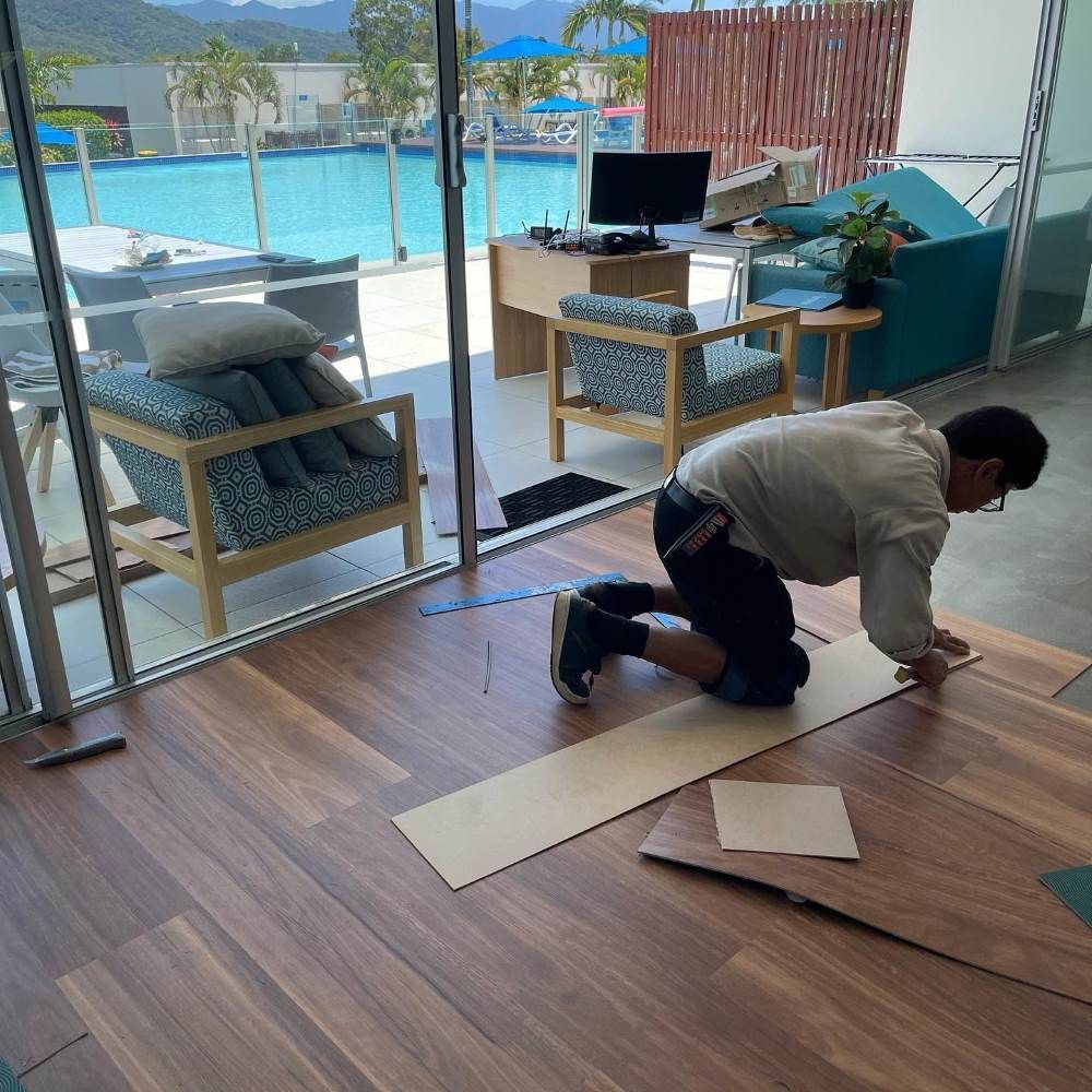 A Man is Kneeling on a Wooden Floor in Front of a Pool — John Chisholm Flooring in Manunda, QLD