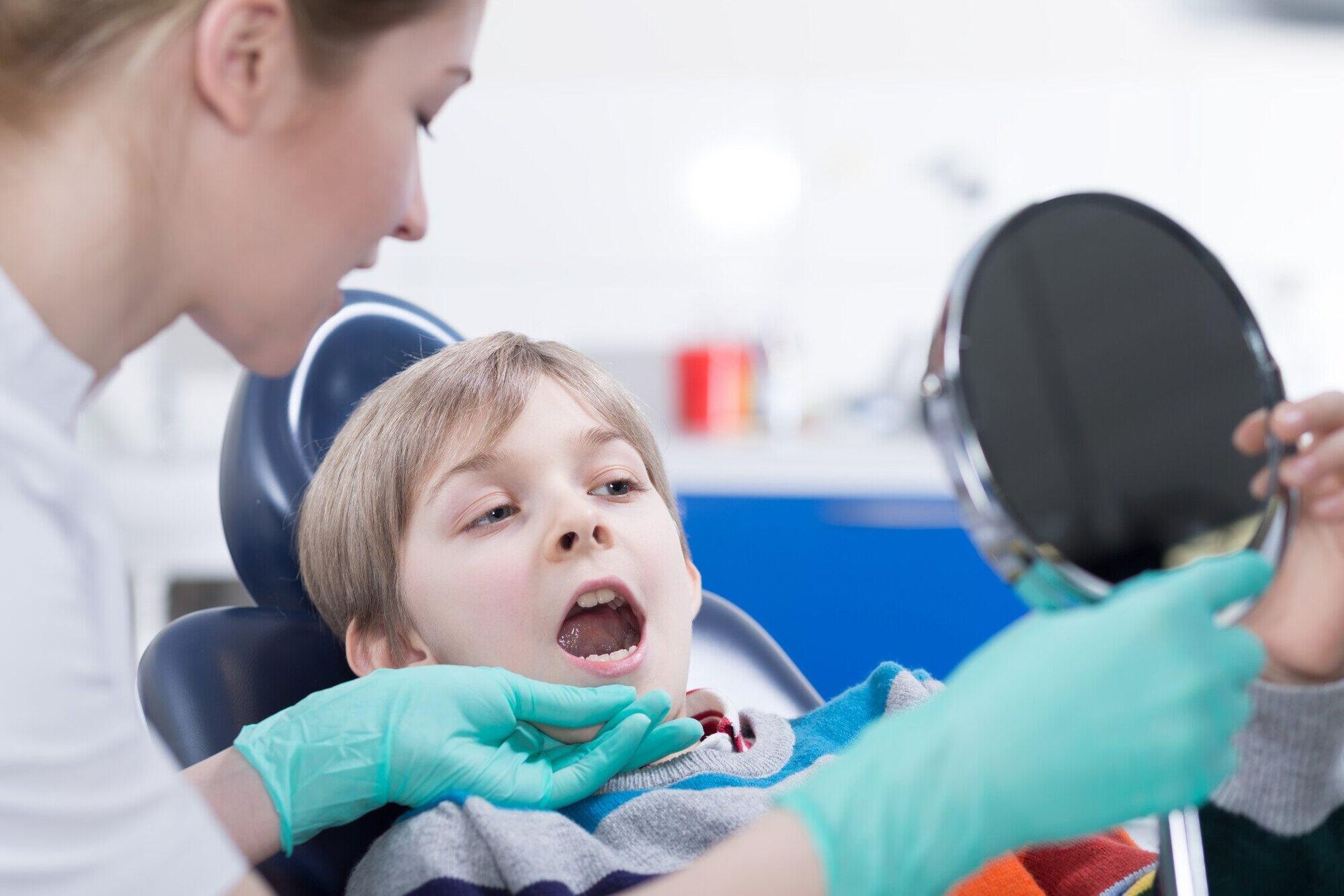 A young boy is sitting in a dental chair looking at his teeth in a mirror.