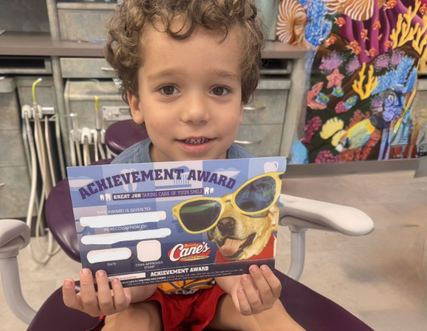 A young child holds a Raising Cane's achievement award while sitting in a dental chair.