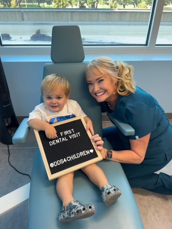 Smiling woman kneels beside child holding a sign that says “First Dental Visit” at a clinic.