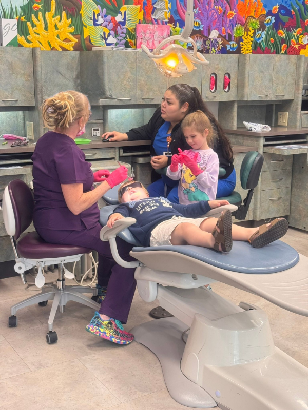 A dental professional examines a patient in a chair, while an assistant and a child wearing gloves look on in a room.