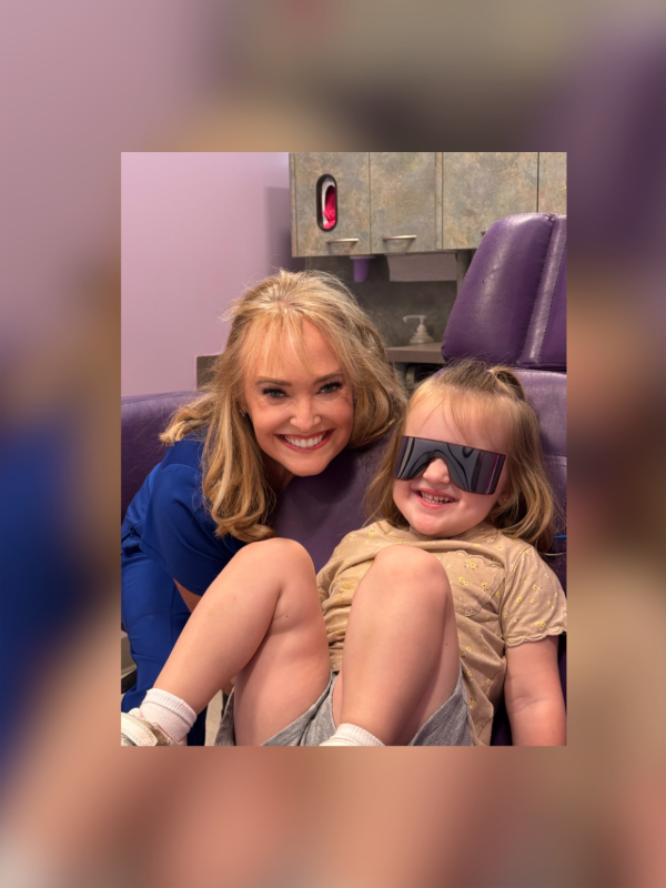 A smiling healthcare professional leans over a young child wearing dark, protective eye shields in a dental-style chair.