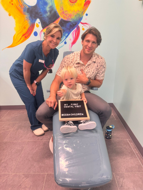 A dental professional stands beside a parent and toddler holding a sign reading