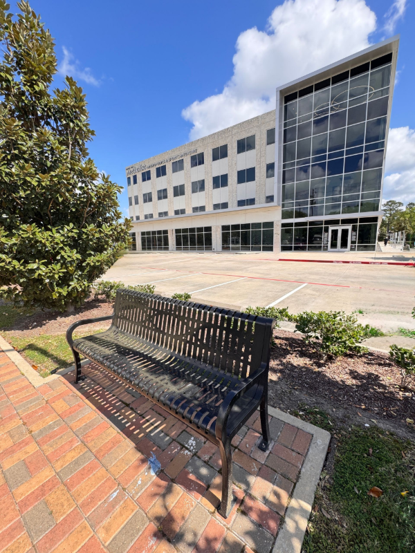 A black metal bench sits on a brick path in front of a modern four-story office building under a blue, cloudy sky.