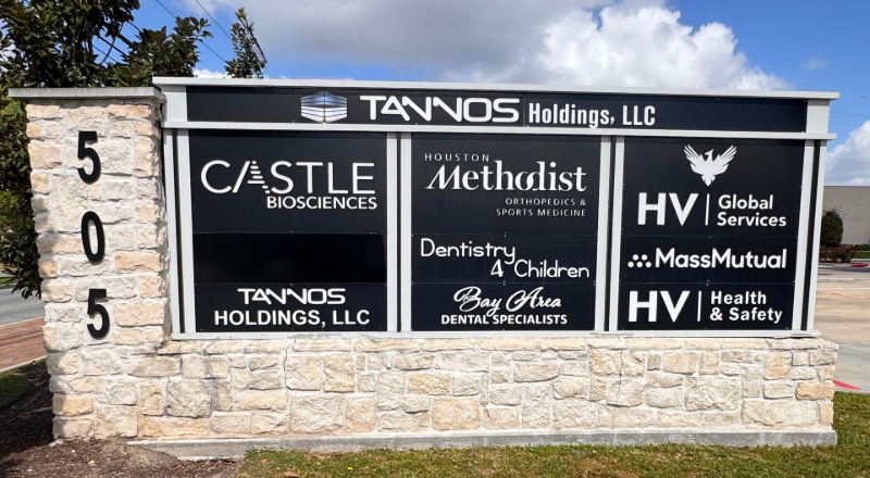 A stone monument sign for Tannos Holdings, LLC at 505 listing multiple businesses on a black panel against a blue sky.