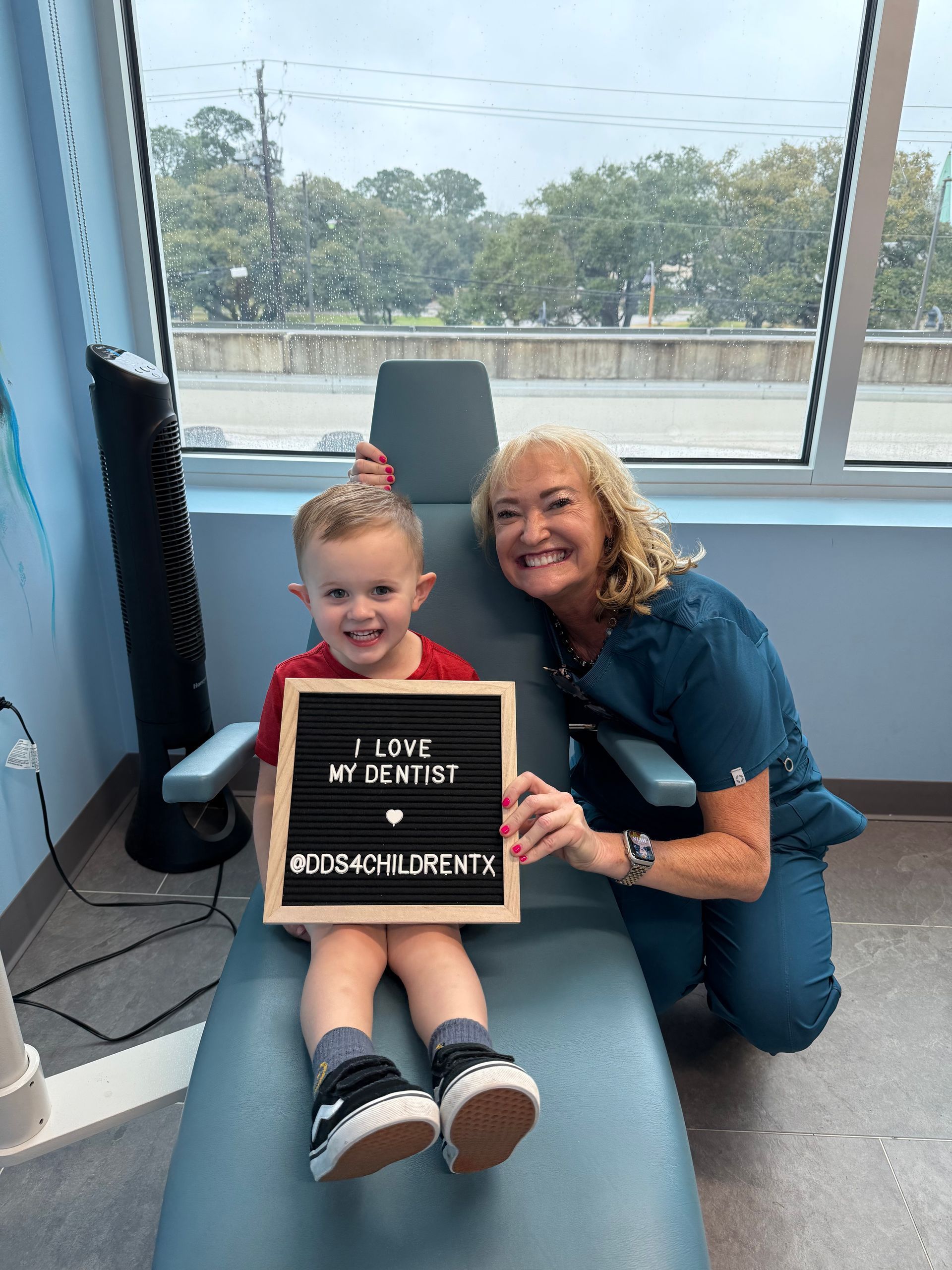 A woman is kneeling next to a young boy in a dental chair holding a sign.