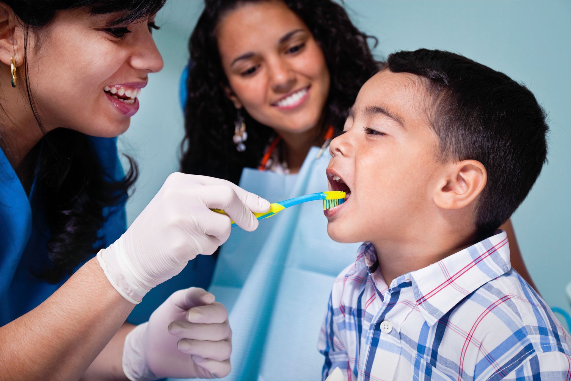 Dental worker wearing gloves brushes a child’s teeth while another woman smiles nearby