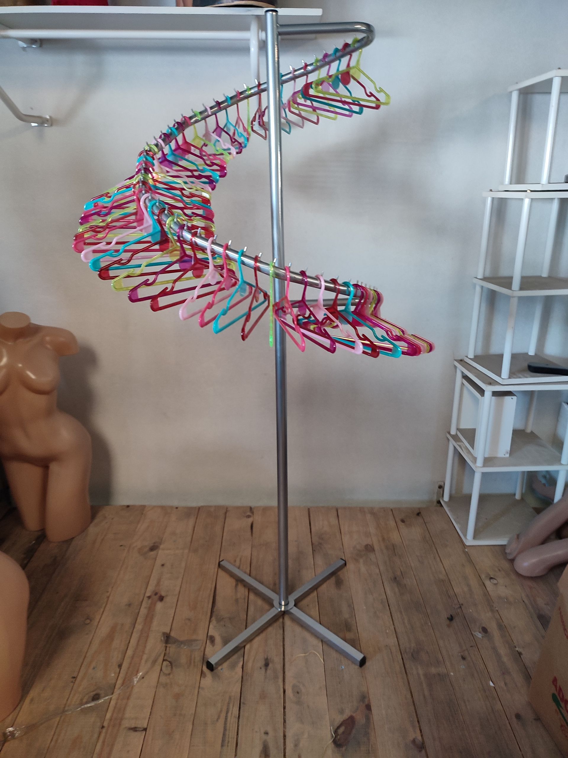 Perchero plateado en espiral con perchas de colores. Suelo de madera y estantes blancos al fondo.