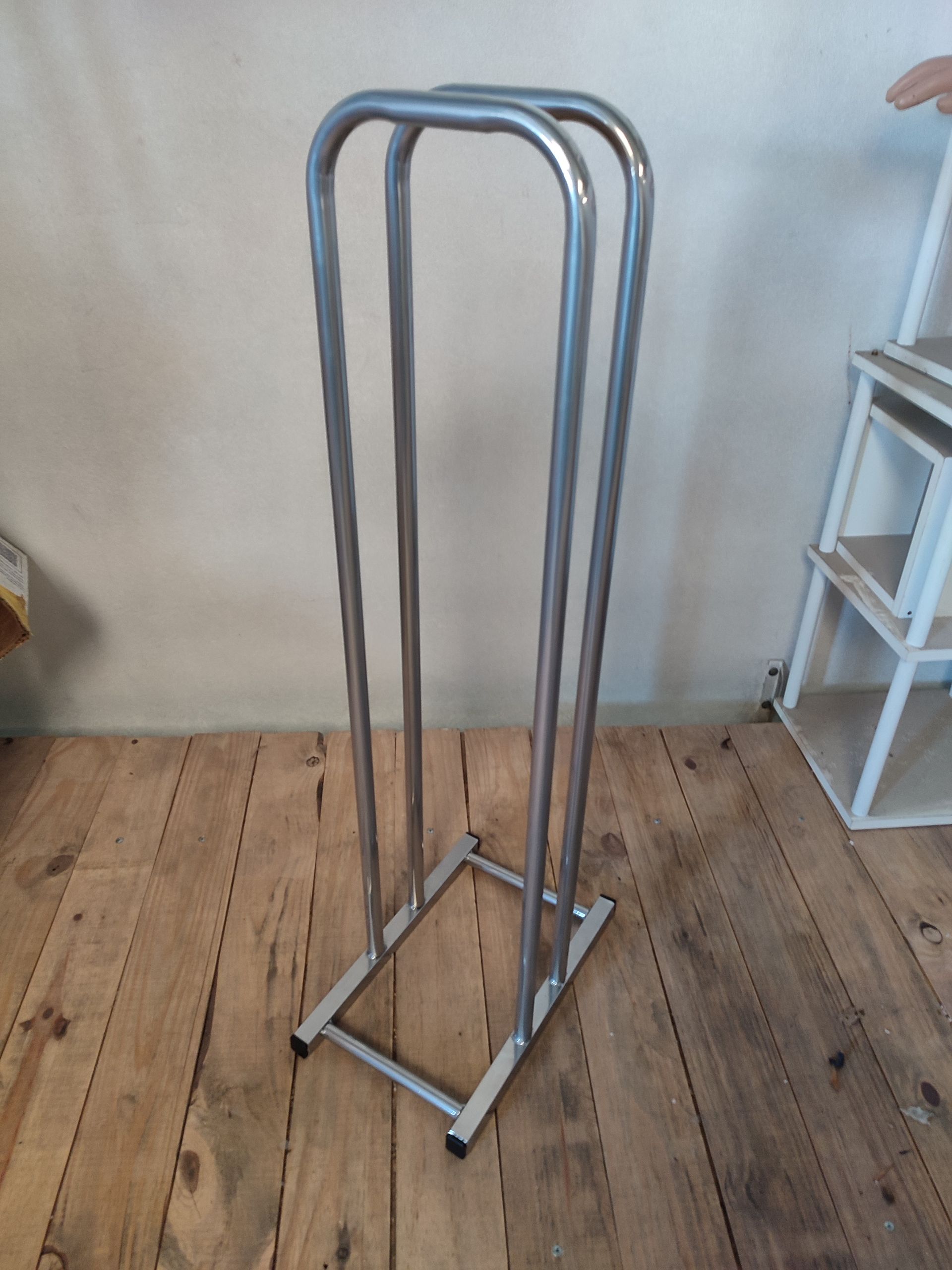 Toallero metálico sobre suelo de madera con pared blanca y estantería al fondo.