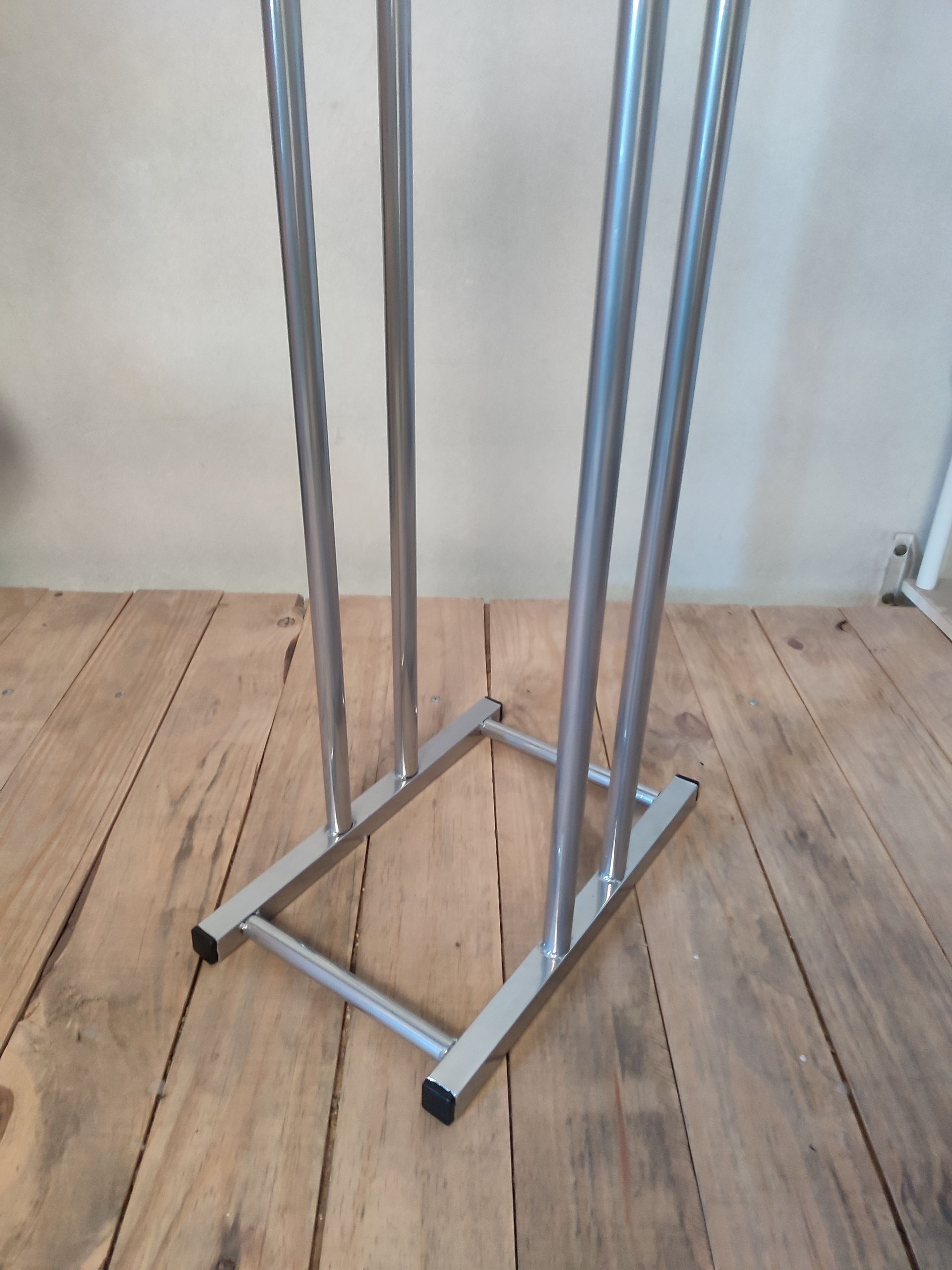 Perchero metálico sobre suelo de madera, visto desde la base. Marco plateado con cuatro barras verticales.