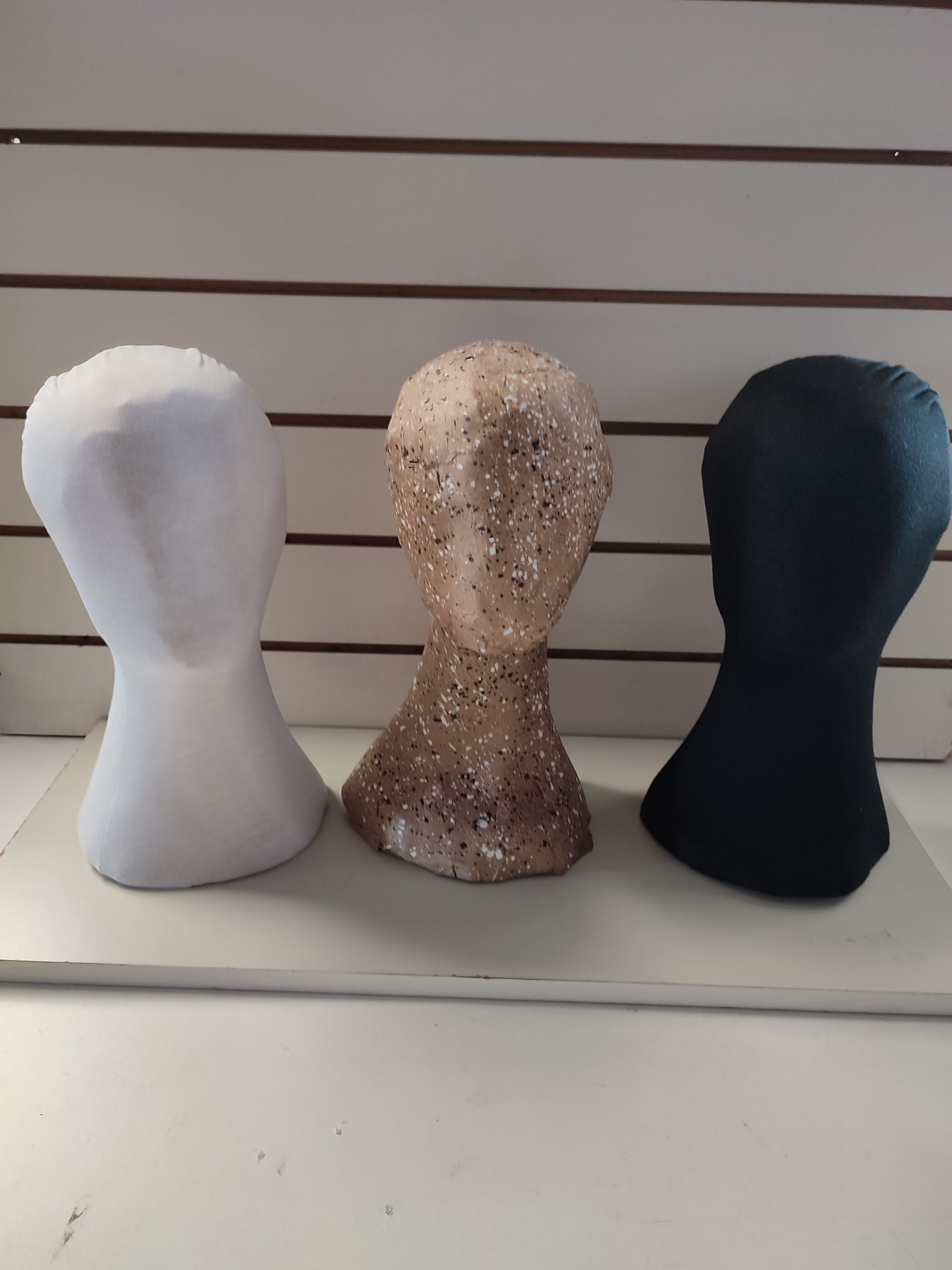Tres cabezas de maniquí: blanca, dorada con brillo y negra, alineadas en un estante.