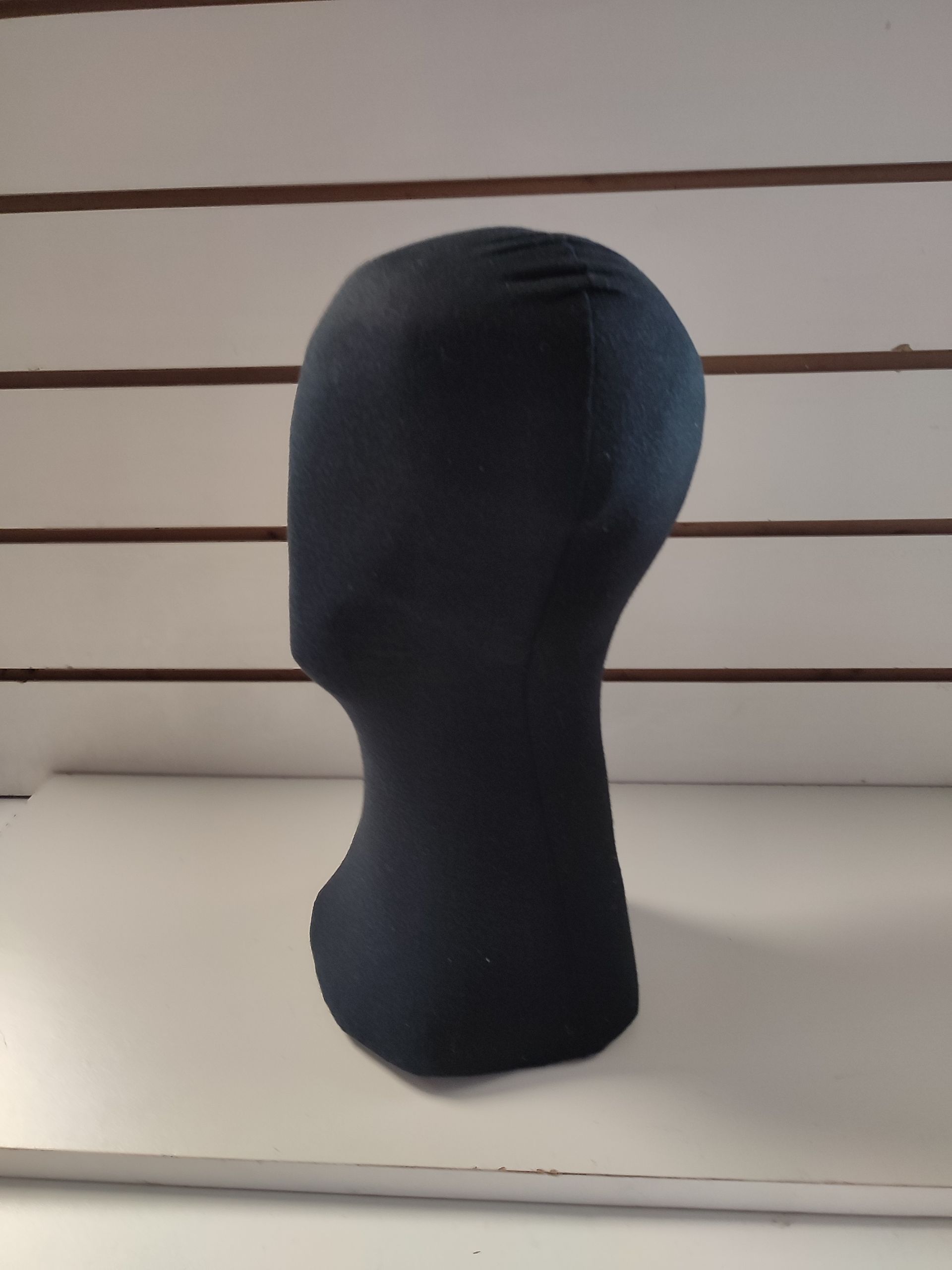 Cabeza y cuello de maniquí negro sobre una superficie blanca, sobre un fondo de listones horizontales marrones.