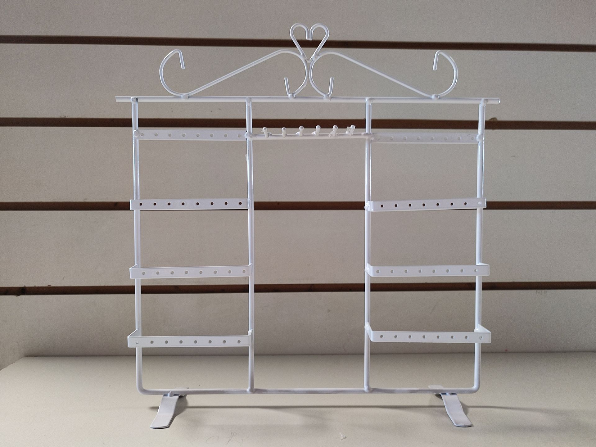 Organizador de joyas de metal blanco con estantes y detalle de corazón, contra una pared revestida de madera.