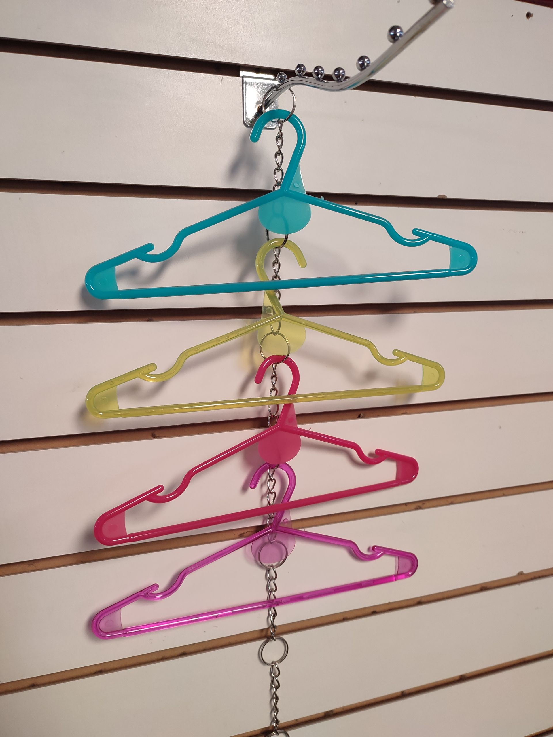 Tres perchas de plástico de colores colgadas verticalmente en una pared ranurada, conectadas por una cadena.