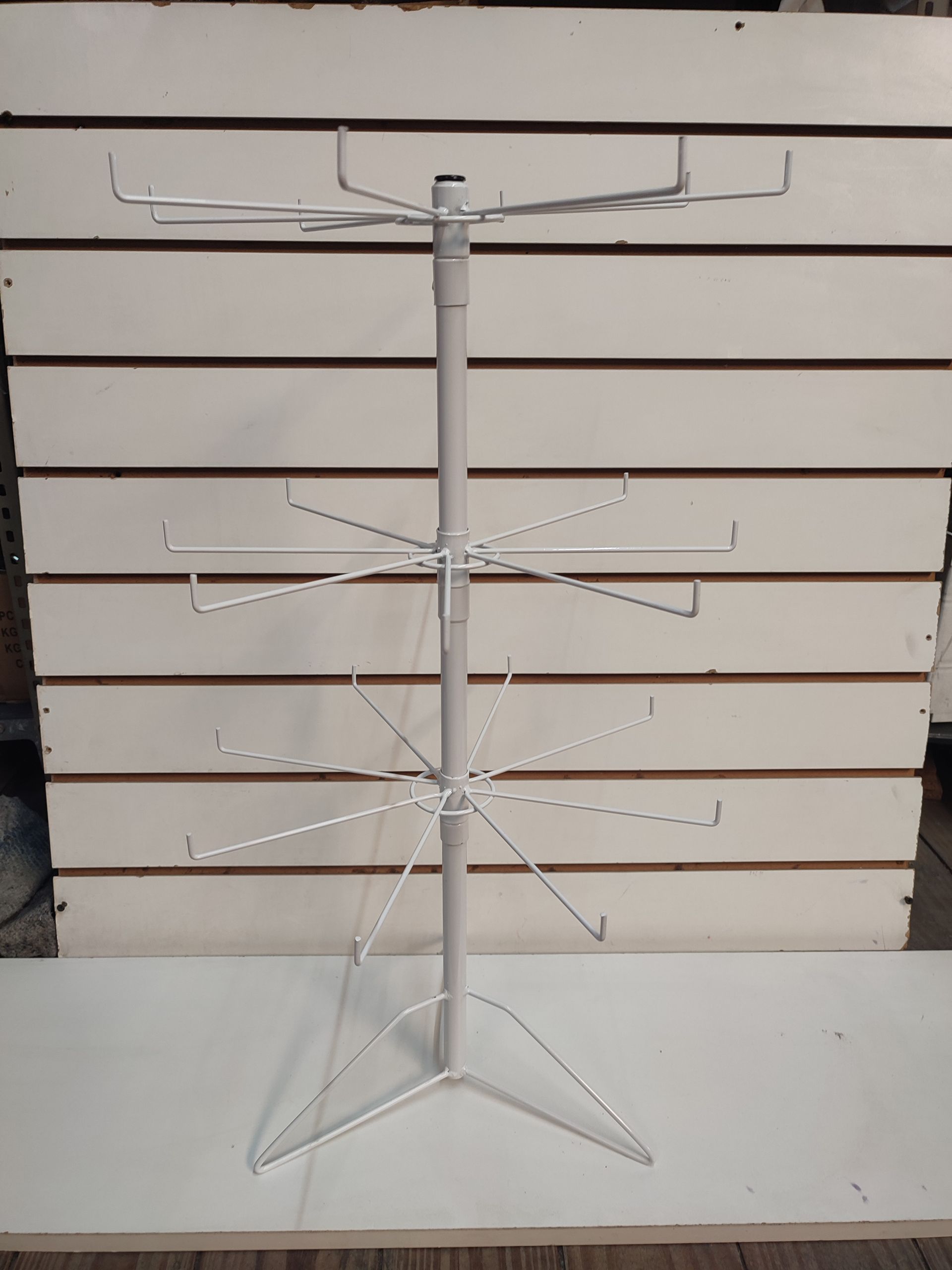 Expositor giratorio blanco con múltiples brazos, sobre una superficie blanca, contra una pared revestida.