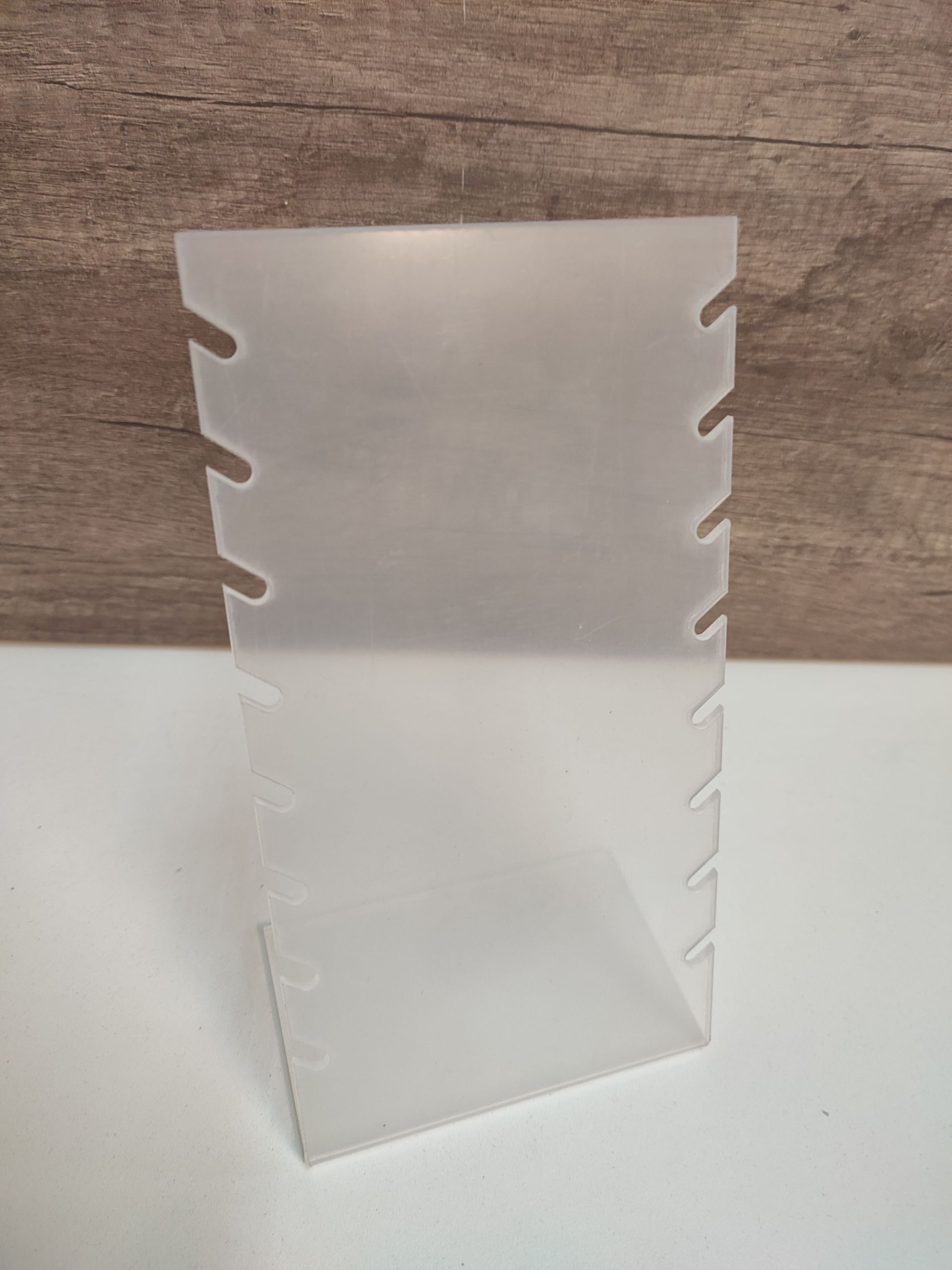 Expositor de joyas de plástico esmerilado con ranuras. Sobre una superficie blanca, con fondo de veta de madera.