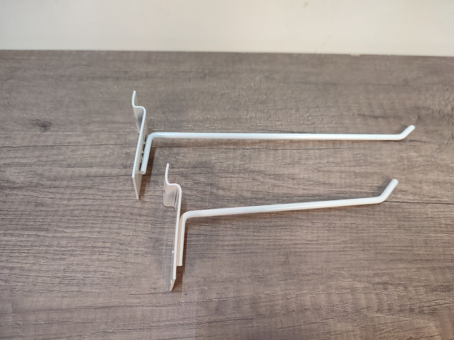 Dos ganchos de exhibición minorista de metal blanco sobre una superficie con vetas de madera.