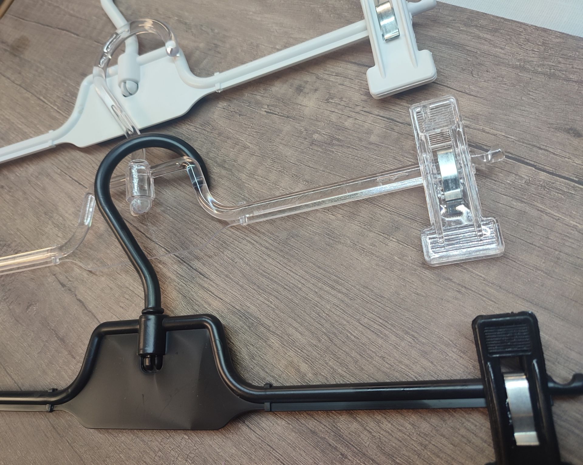 Tres perchas de ropa con clips: de plástico negro, blanco y transparente, sobre una superficie con vetas de madera.