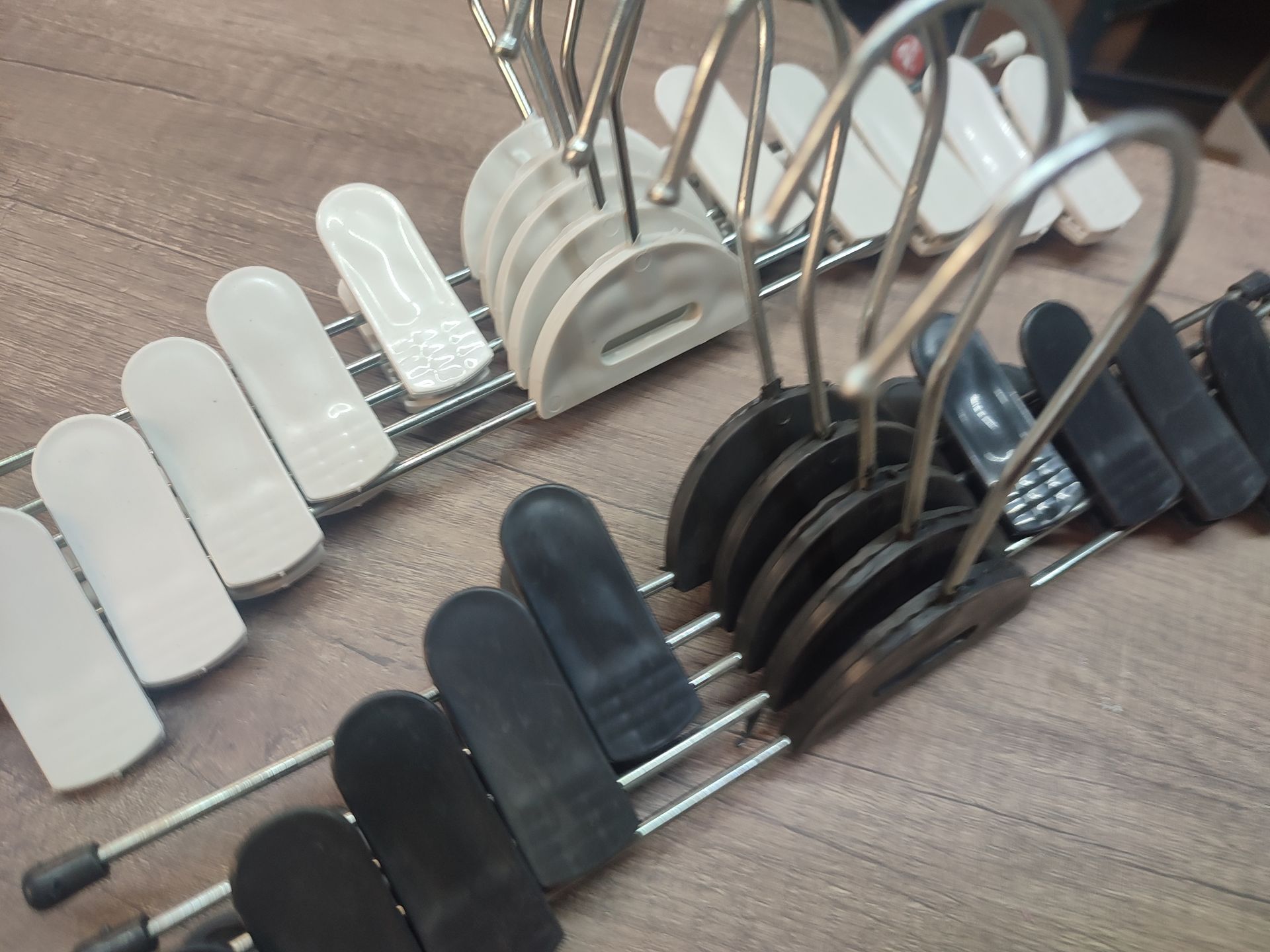 Varias perchas para pantalones, algunas de color negro, blanco y tostado, con pinzas sobre una superficie de madera.