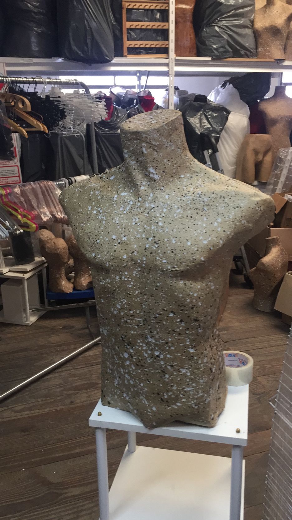 Torso de maniquí con superficie texturizada, sobre un soporte blanco en un almacén desordenado.