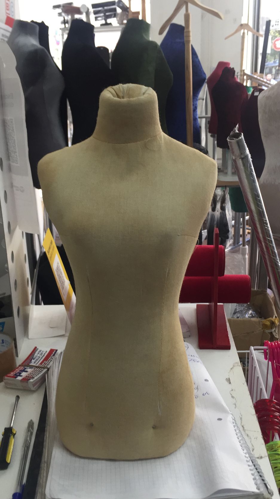 Torso de maniquí de modista en tela color canela sobre una mesa con herramientas y otros maniquíes al fondo.