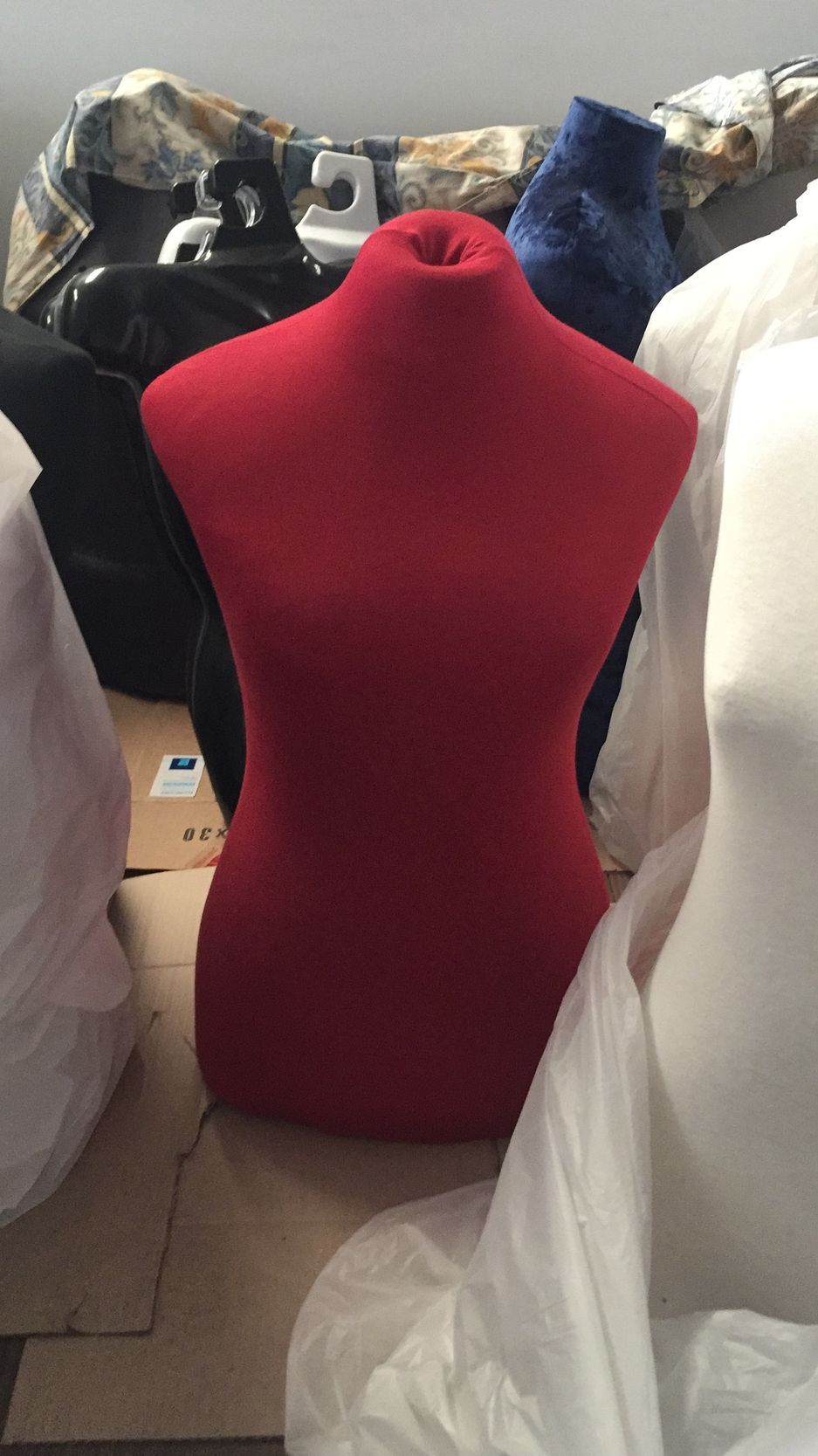 Figura de modista de color rojo, solo el torso, rodeada de otras formas y telas almacenadas.