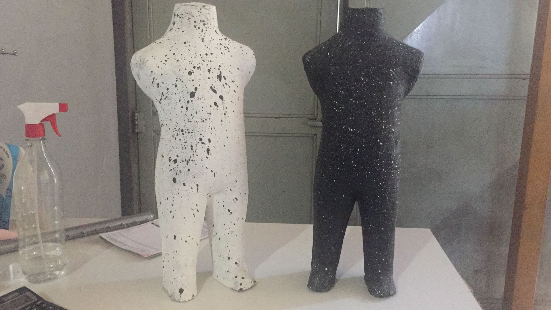 Dos maniquíes sin cabeza, uno blanco y otro negro, salpicados de pintura contrastante, están de pie sobre una mesa.