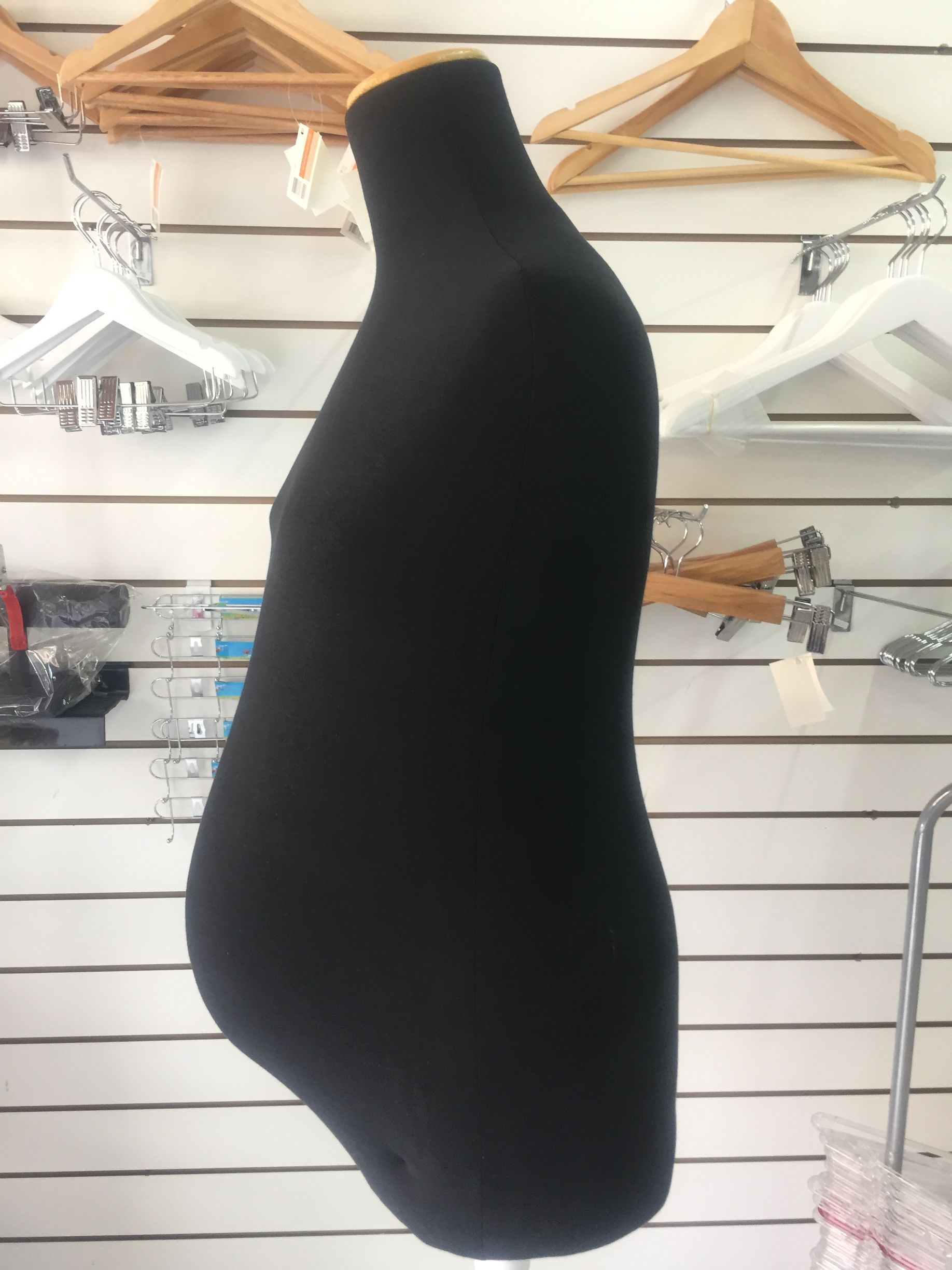 Maniquí de vestir negro, vista lateral, sobre una pared de listones con varias perchas.