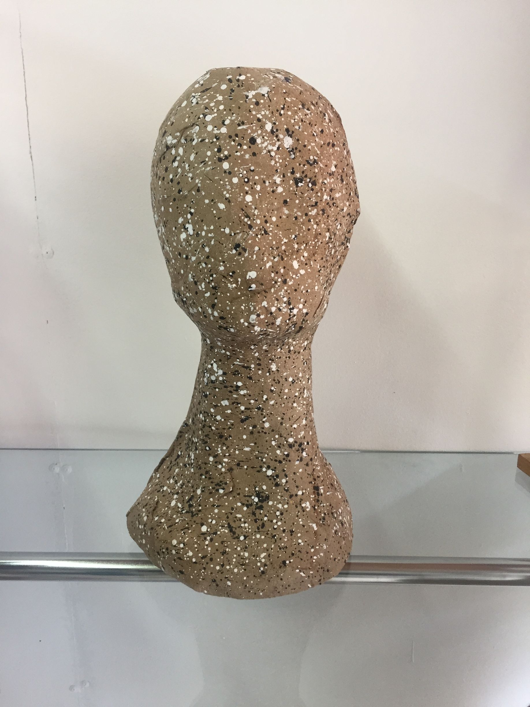 Escultura en forma de cabeza de color marrón con motas blancas y negras sobre un estante blanco.
