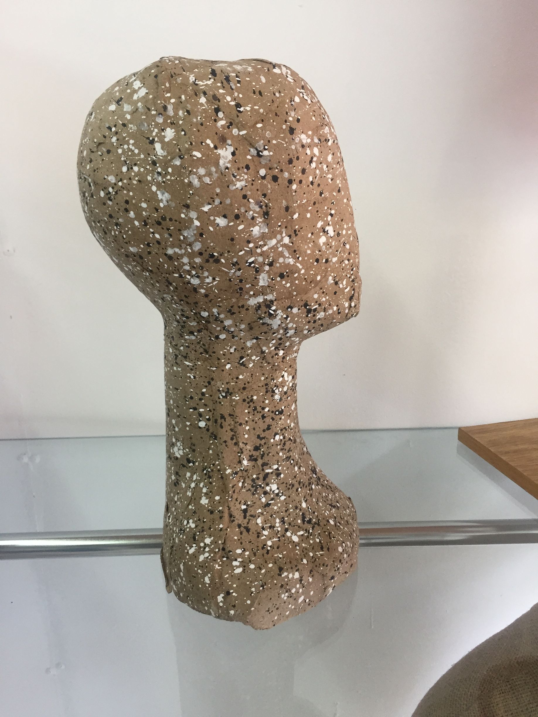 Cabeza y cuello de maniquí, de color canela moteado con motas blancas y negras, sobre un estante.