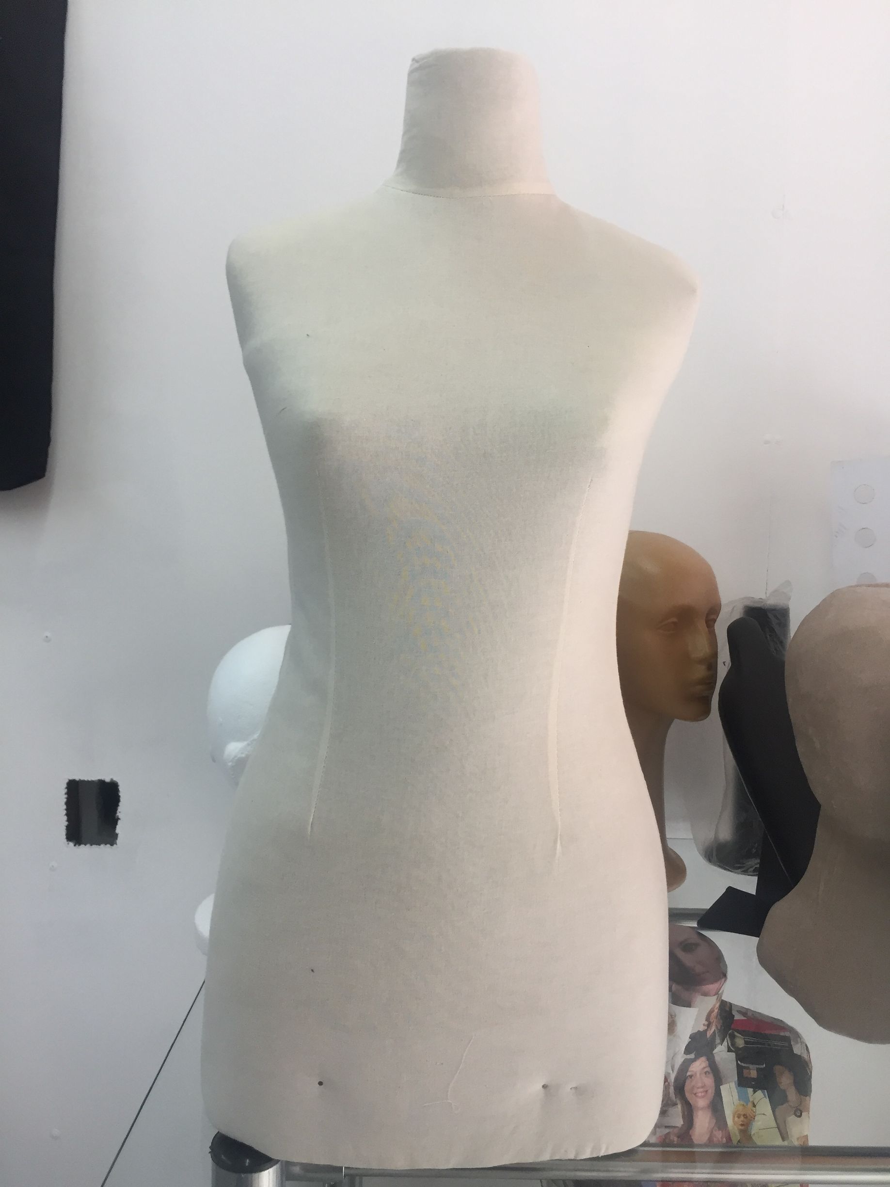 Figura de modista de color crema con una ligera forma de reloj de arena, situada delante de varias cabezas de maniquí.