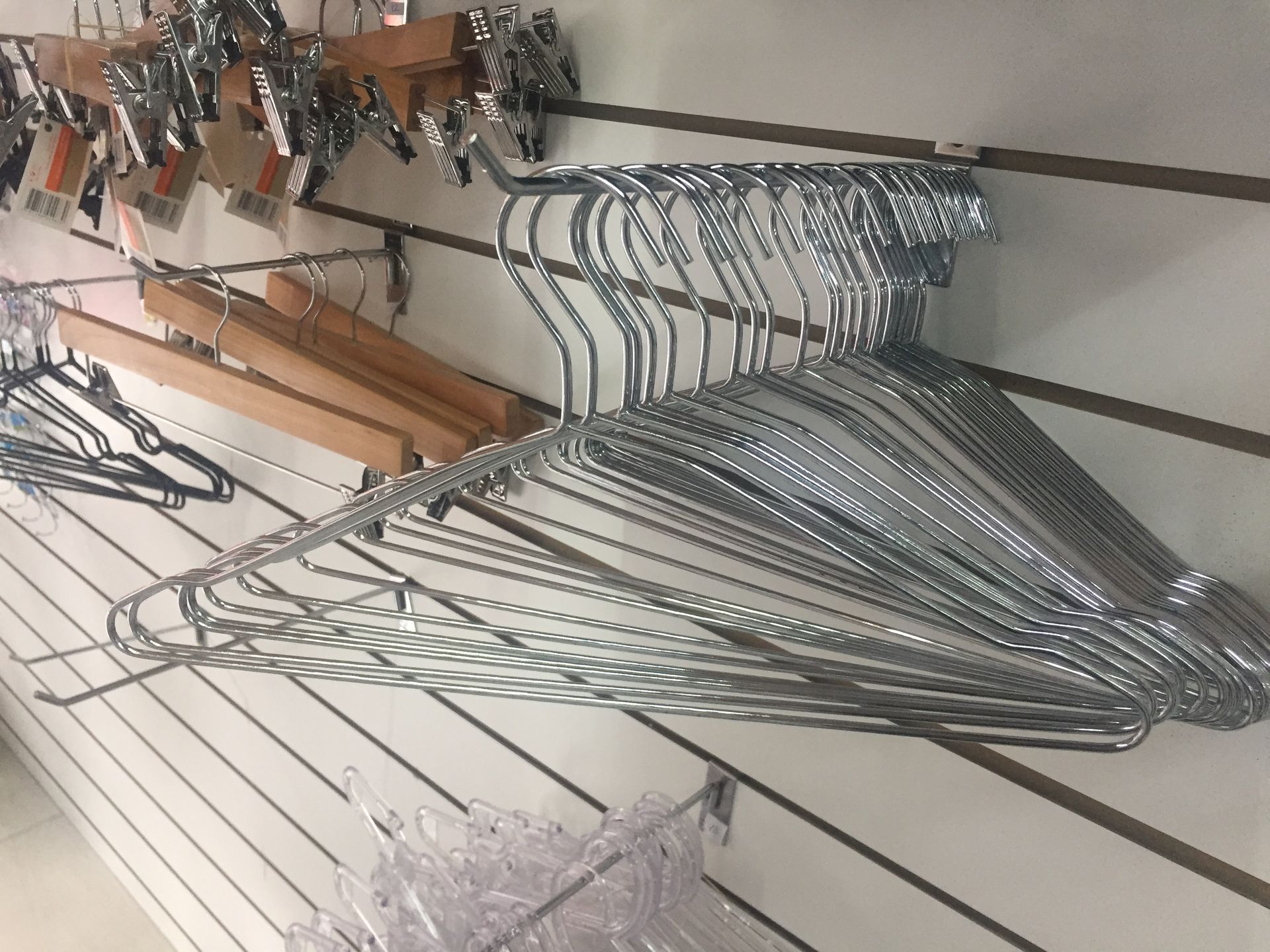 Perchas de metal expuestas en una pared ranurada, junto a perchas de madera y de plástico transparente.