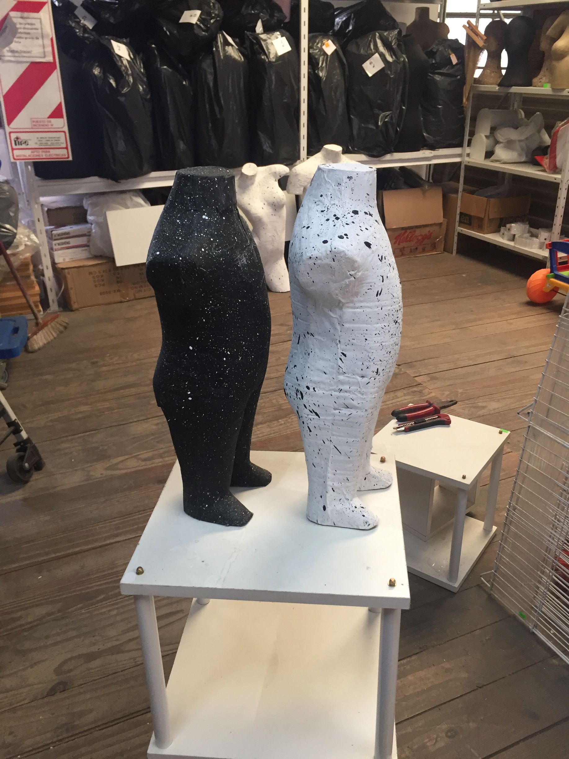 Tres torsos de maniquí, con manchas blancas y negras, se encuentran sobre un estante blanco. Ropa oscura cuelga al fondo.