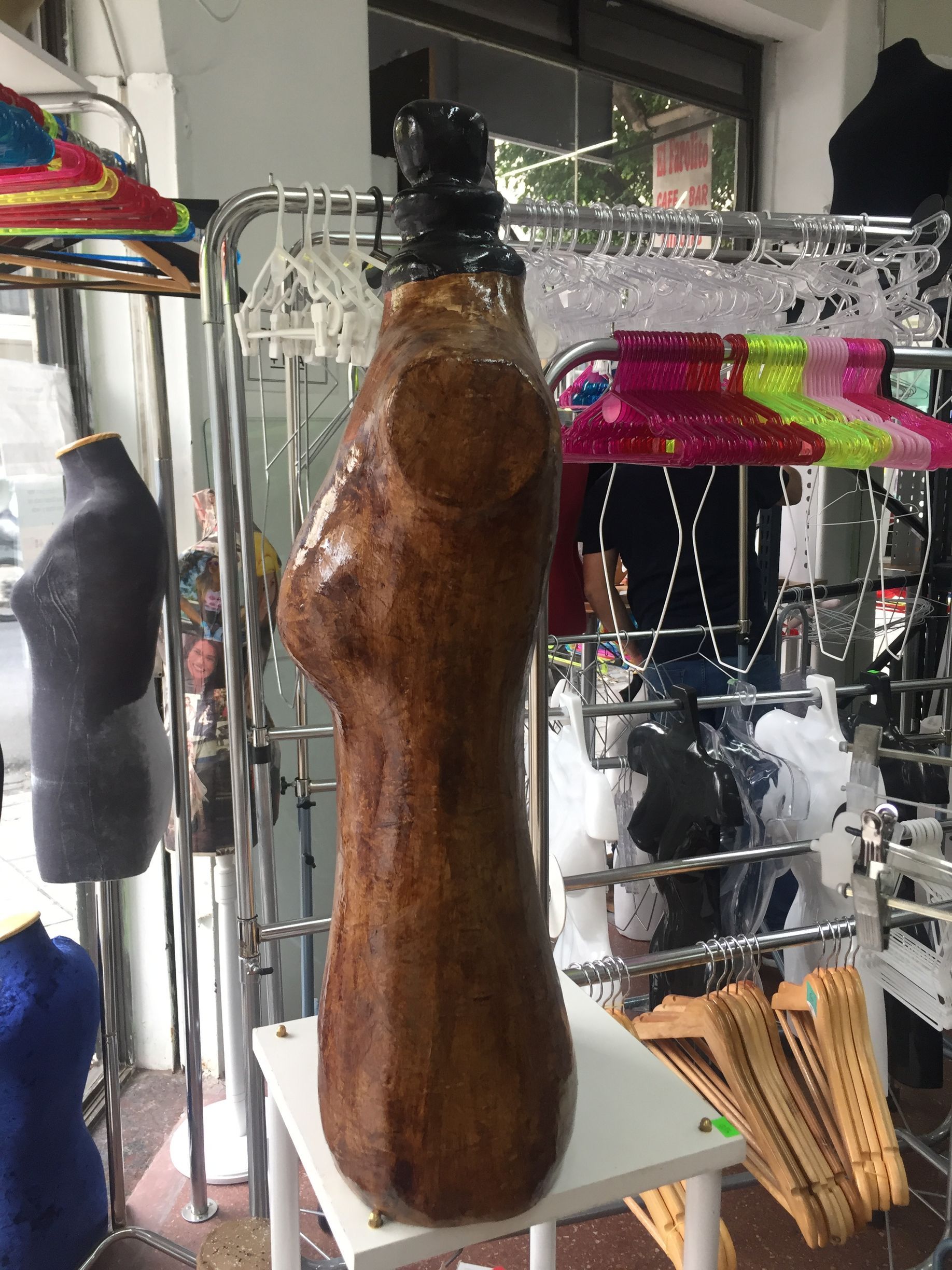 Maniquí de torso de madera sobre una mesa blanca en una tienda de ropa, perchas en el fondo.