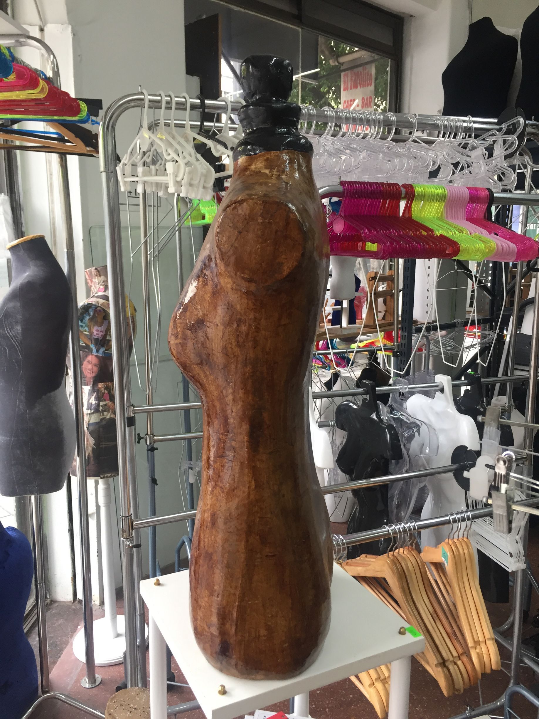 Maniquí de torso marrón con cabeza negra, de pie sobre un estante blanco. Entorno de tienda.