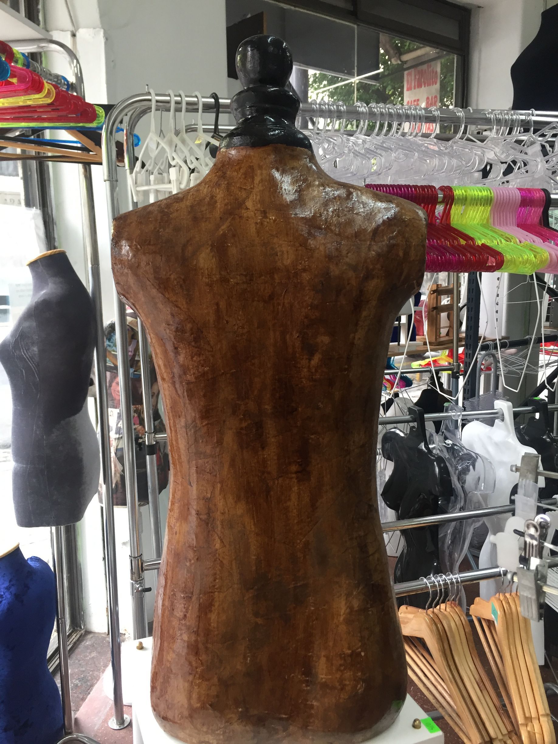 Torso de maniquí marrón en una tienda, con otros maniquíes y ropa visibles.