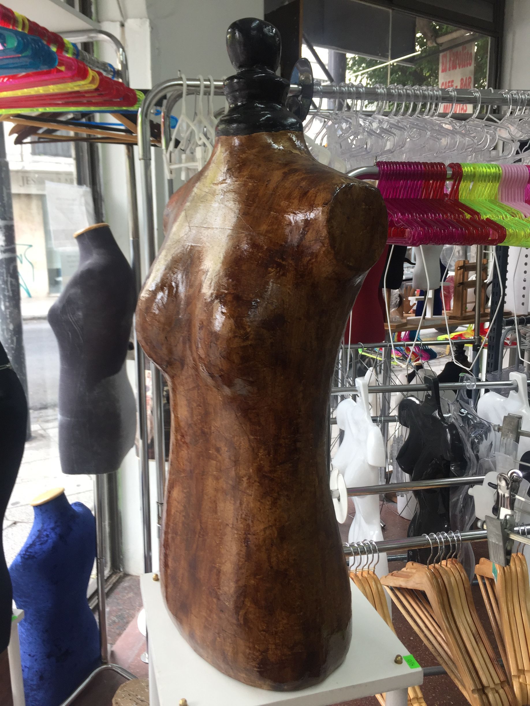 Maniquí de torso de modista de color marrón dentro de una tienda con otros maniquíes y expositores al fondo.